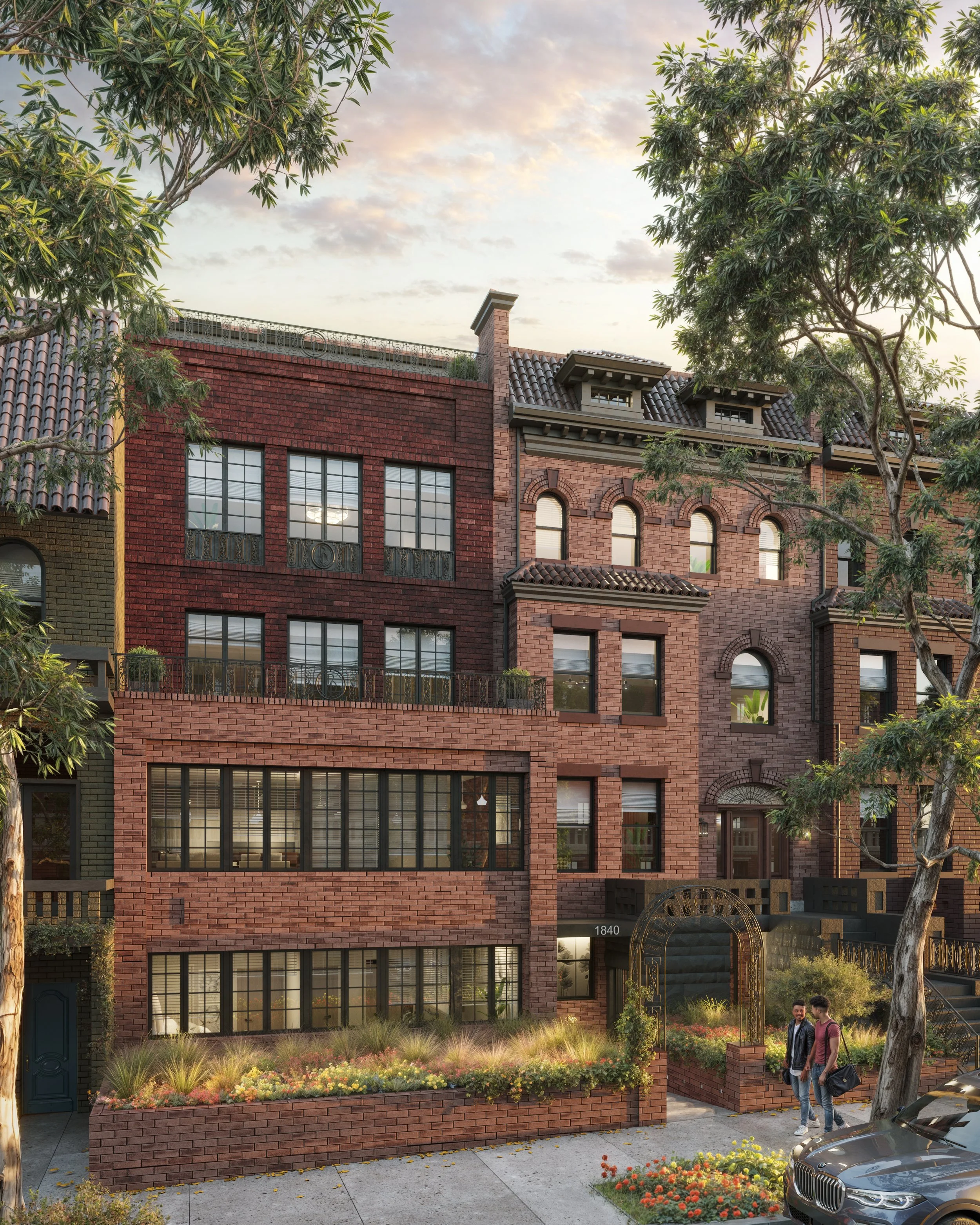 A multi-story brick building with several windows and balconies, surrounded by trees and flowers, at sunset. Two people are walking on the sidewalk near the building, and a car is parked in the street.