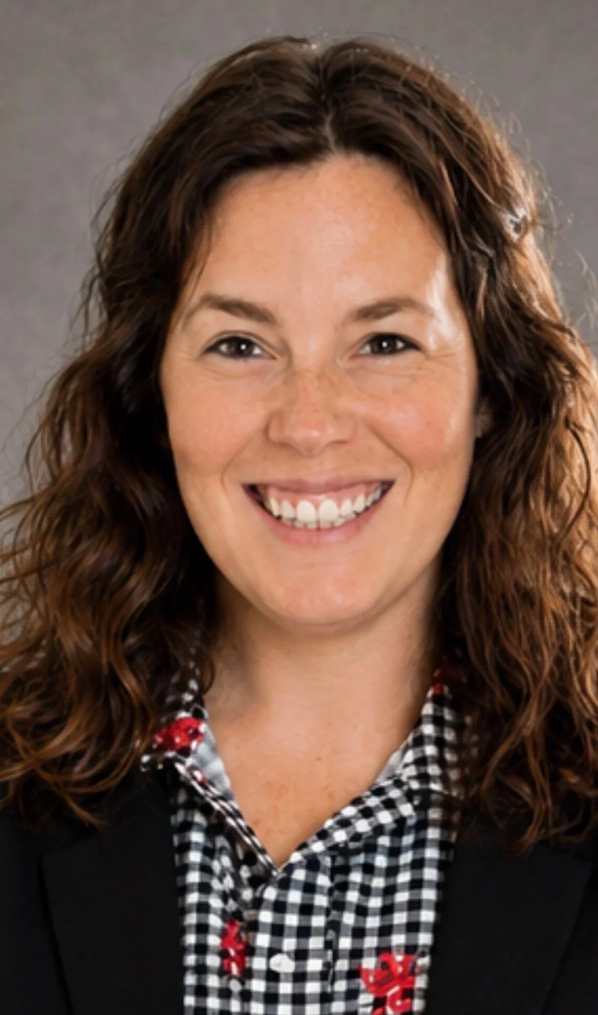 A woman with curly brown hair smiling, wearing a black blazer over a black-and-white checkered shirt with red embroidery.