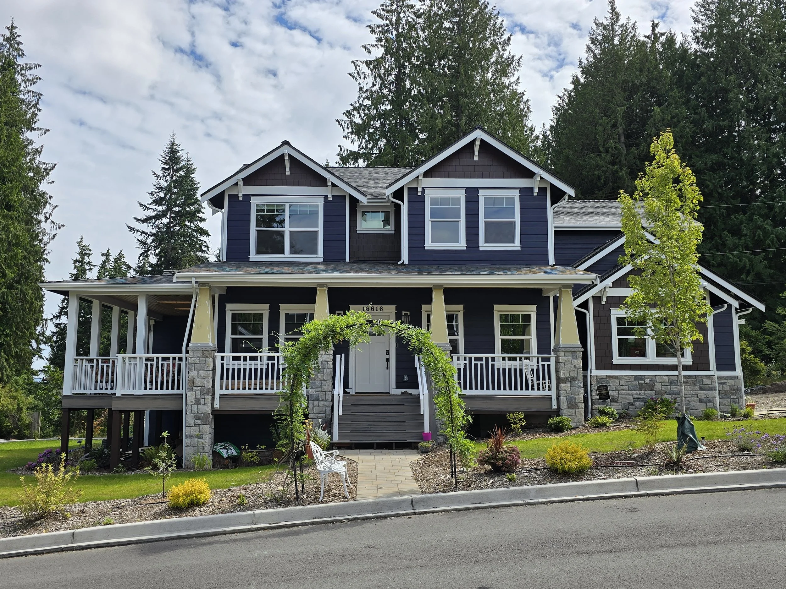 A two-story house painted dark blue with white trim, surrounded by trees and a garden. The house features large windows, a covered porch, and stone pillars. There is a paved walkway leading to the front door and a lawn with various plants and flowers.