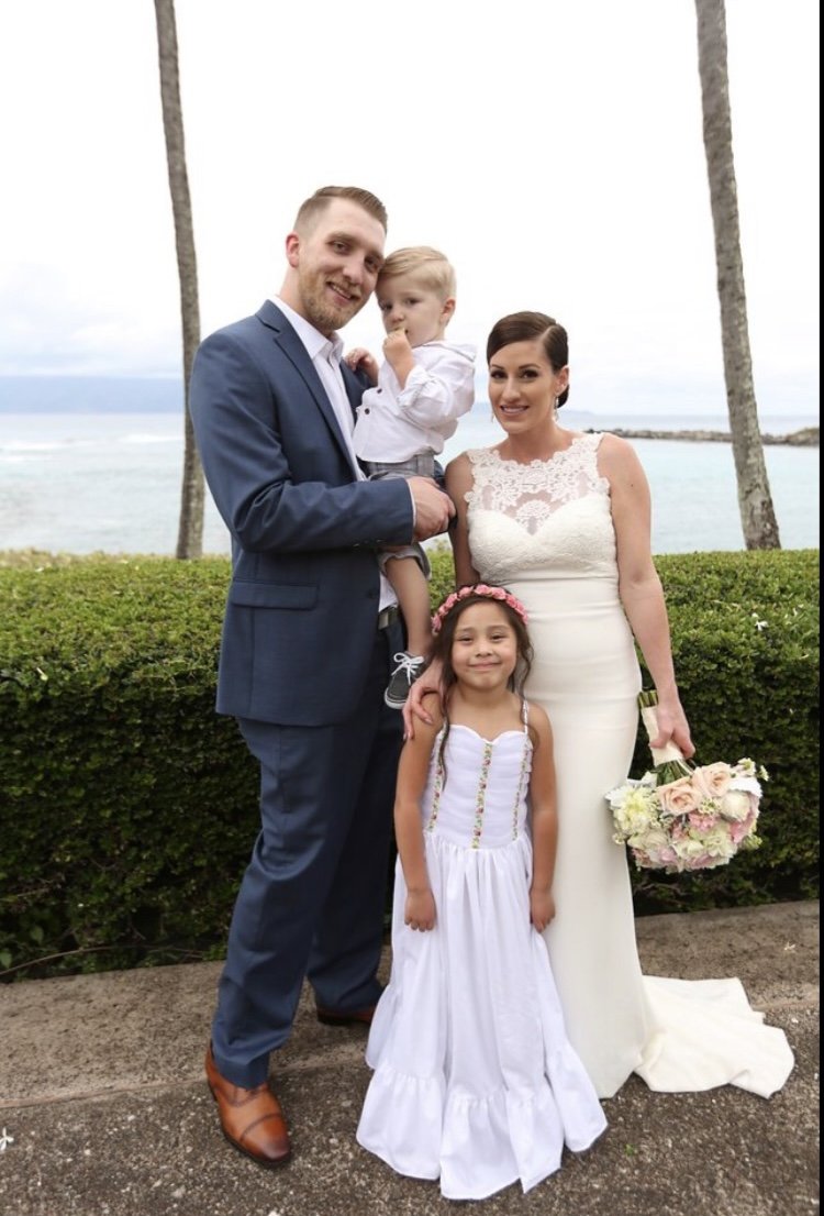 A family of four at a beachside wedding, with the man in a blue suit, the woman in a white wedding dress holding a bouquet, a young girl in a white dress with a flower crown, and a little boy in a white shirt and shorts, standing between palm trees with ocean in the background.