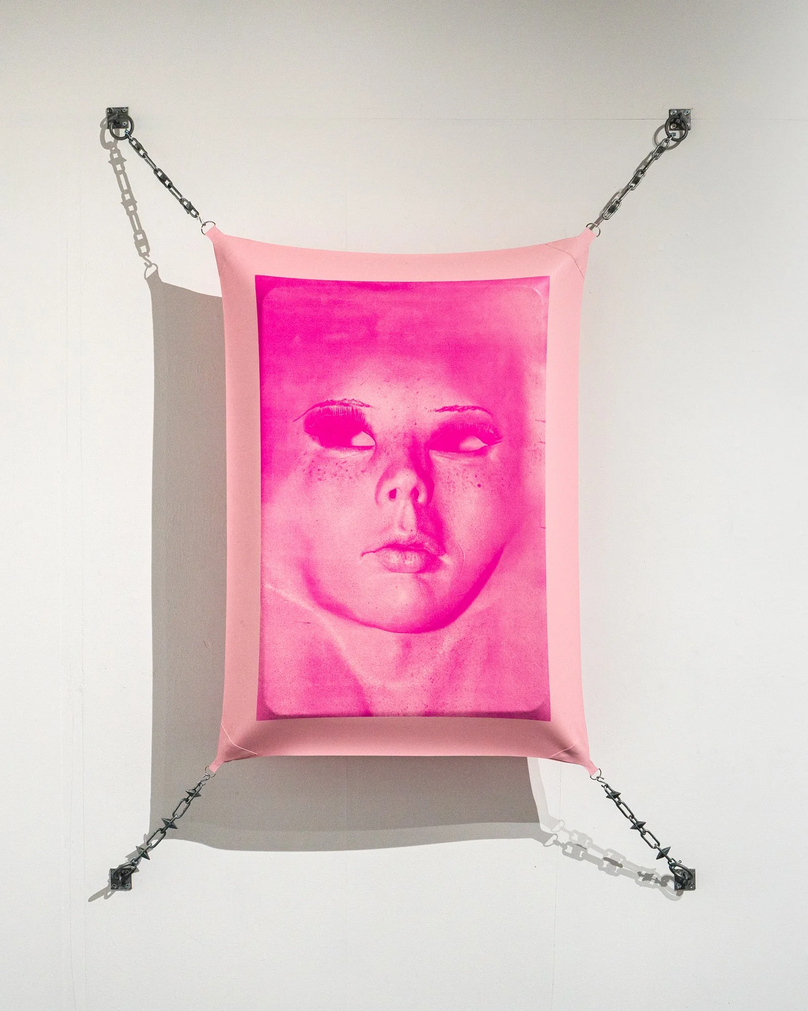 An art installation featuring a large pink fabric with a printed portrait of a woman's face. The fabric is attached to a black metal chain framework on a white wall, creating a square shape with shadows cast on the wall.