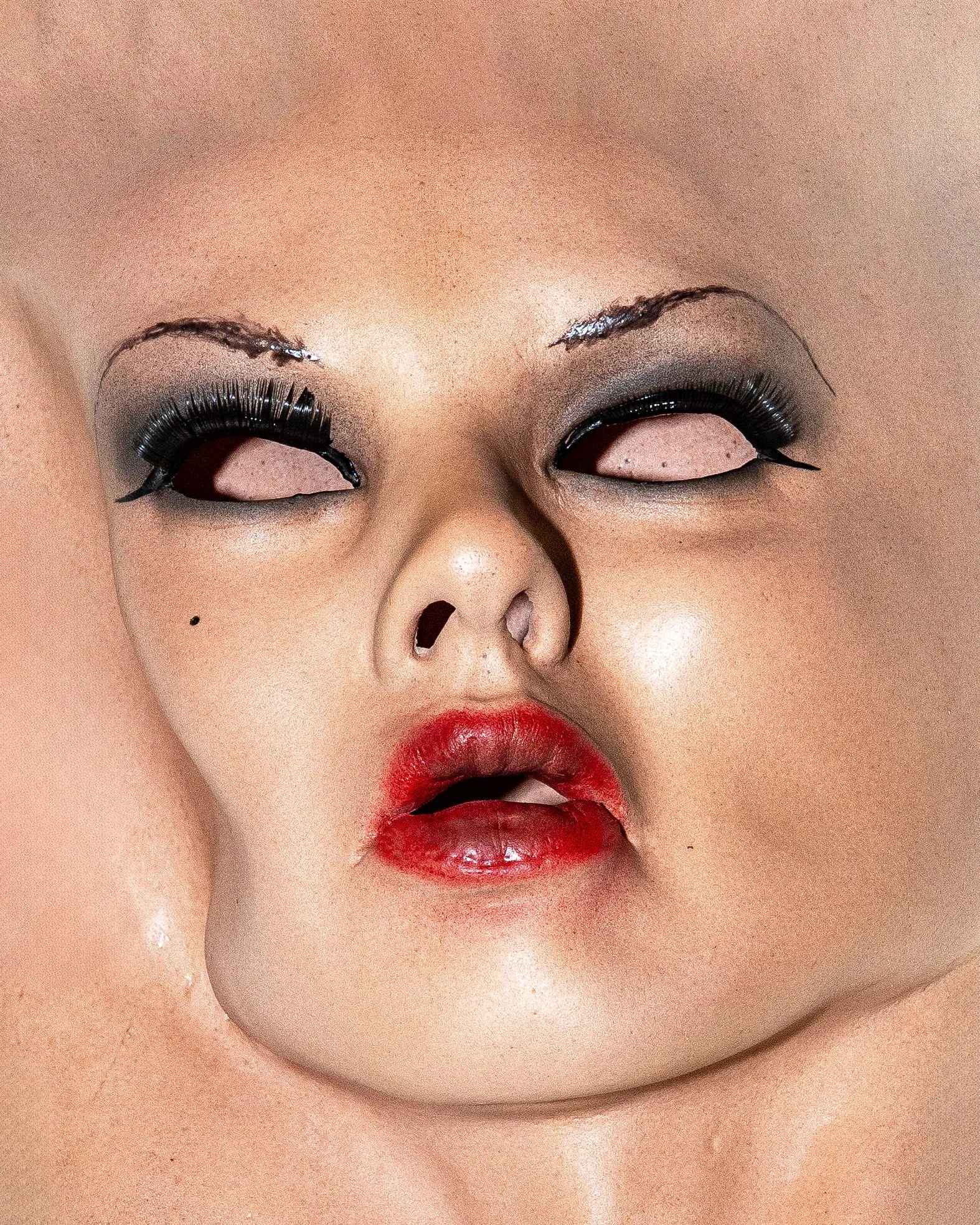 Close-up of a person's face with exaggerated makeup including metallic eye shadow, thick eyeliner, false eyelashes, bright red lipstick, and a small black beauty mark above the lip.