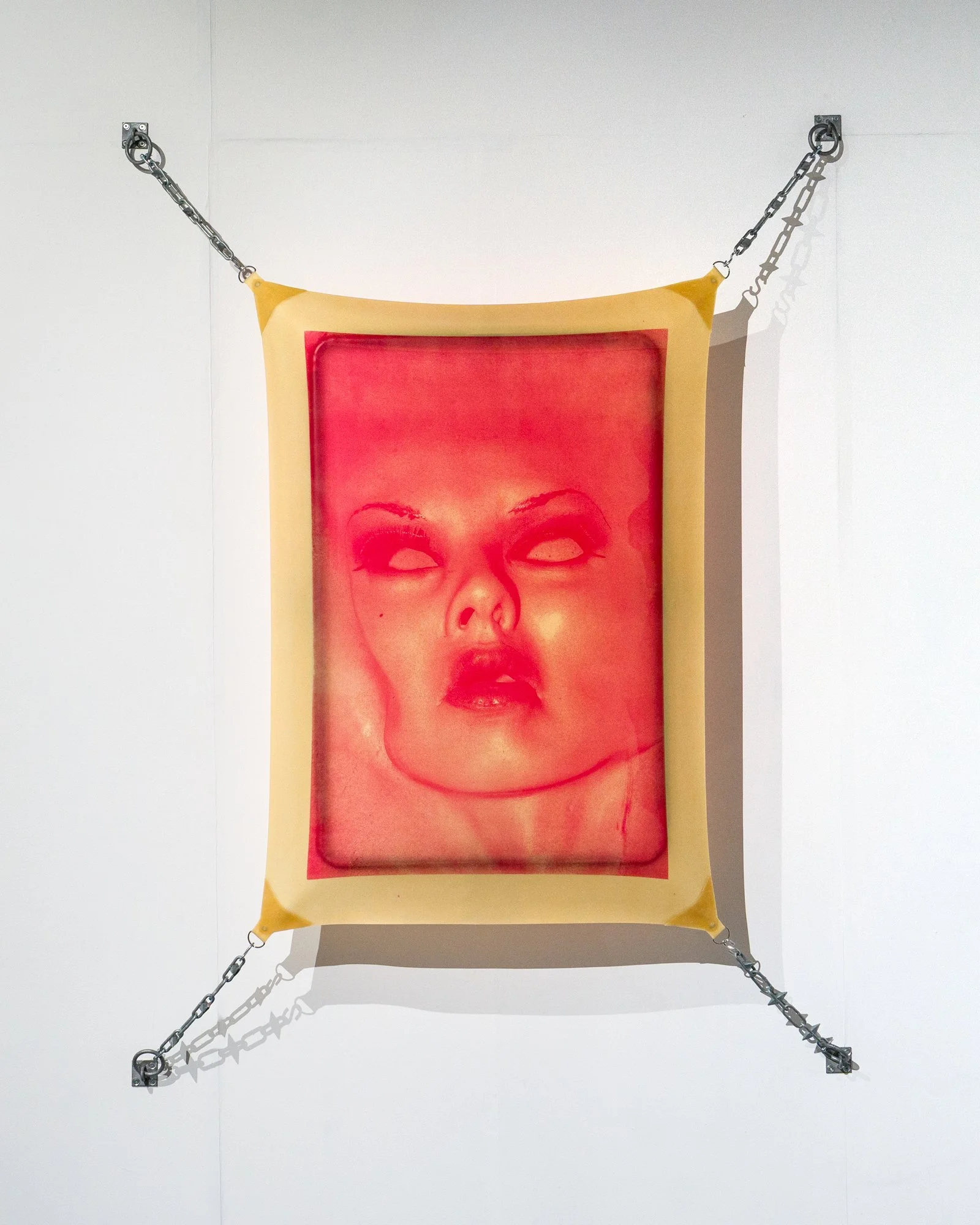 A blank canvas with yellow borders, featuring a portrait of a woman's face in red tones, mounted on a white wall with chains and metal mounted corners.