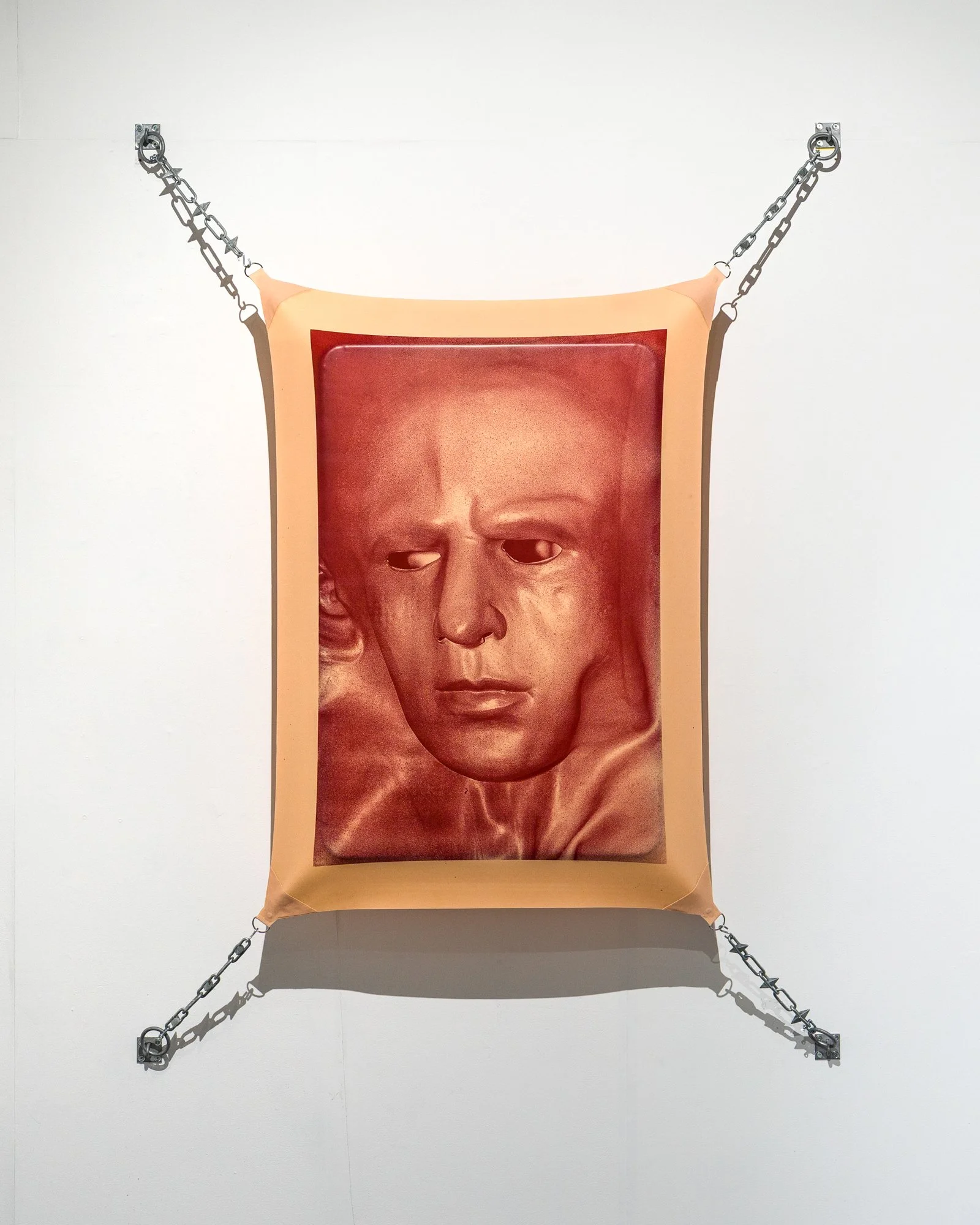 Artwork featuring a large, digitally manipulated portrait of a person's face with a neutral expression, hung on a white wall and secured with metal chains and brackets at the corners.