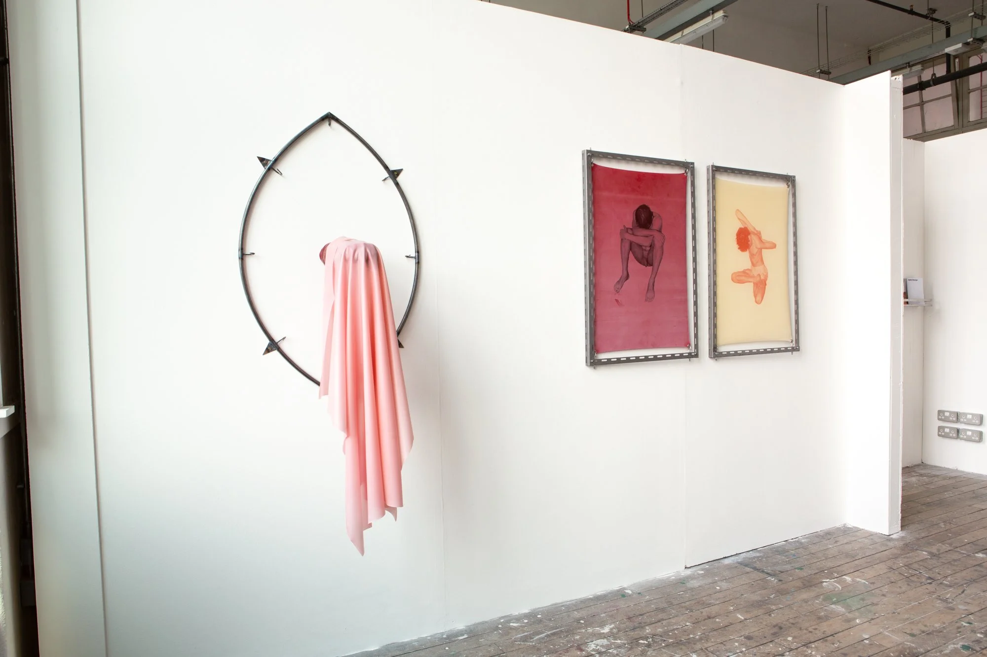 Art installation with two framed pictures of nude women, a pink sculpture of a woman’s head with a pink fabric draped below, and a circular metal frame with pink fabric hanging from it, displayed on a white gallery wall.
