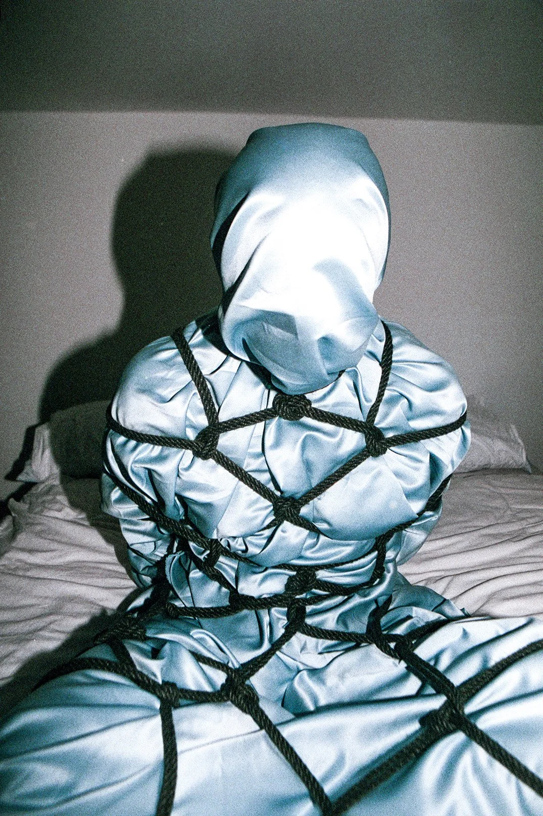 Person in silver protective suit with a hood, bound with black rope, sitting on a bed in a dimly lit room.