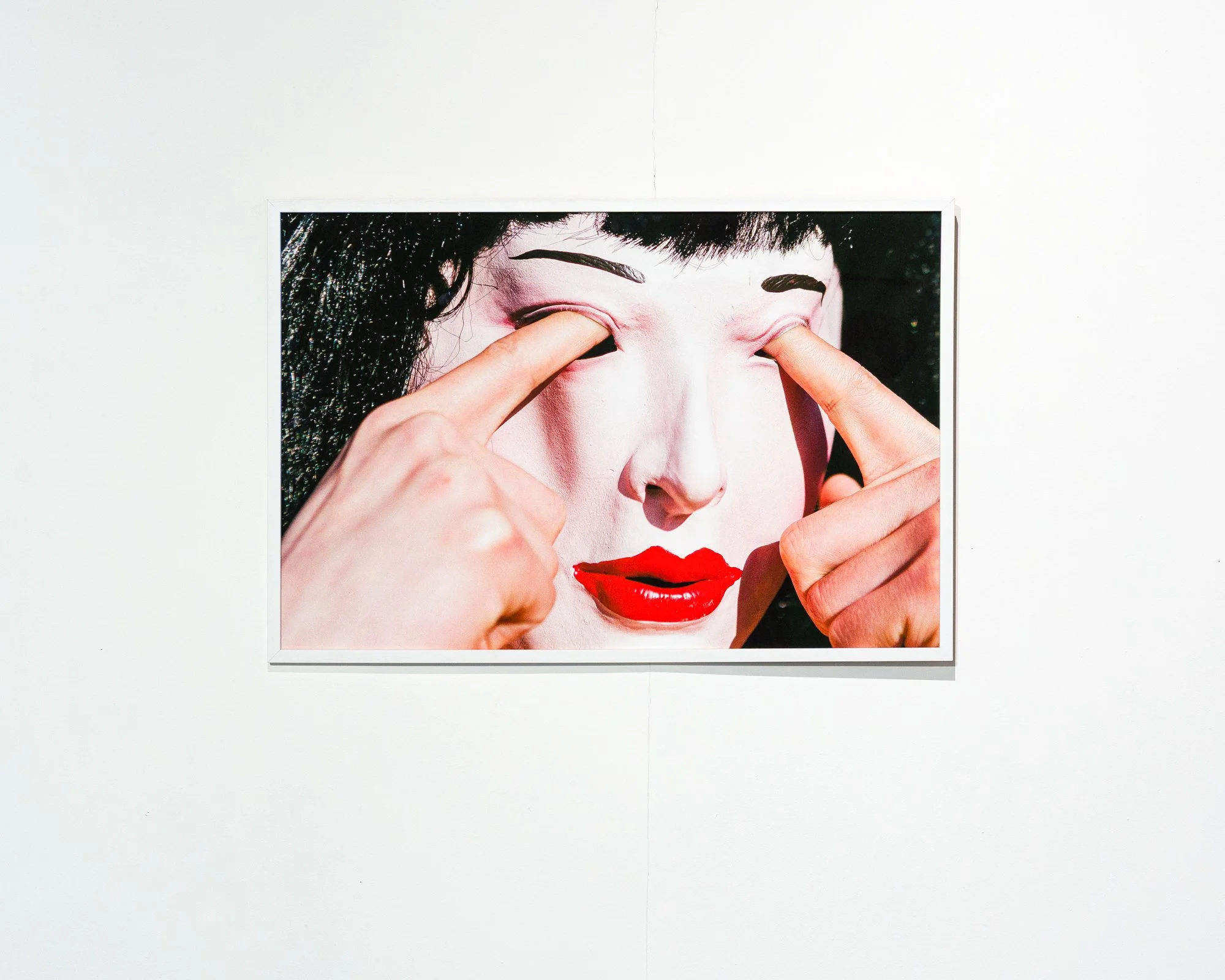 Close-up of a framed art piece showing a woman with black hair, red lipstick, and a light face, touching her eyelids with her fingers.