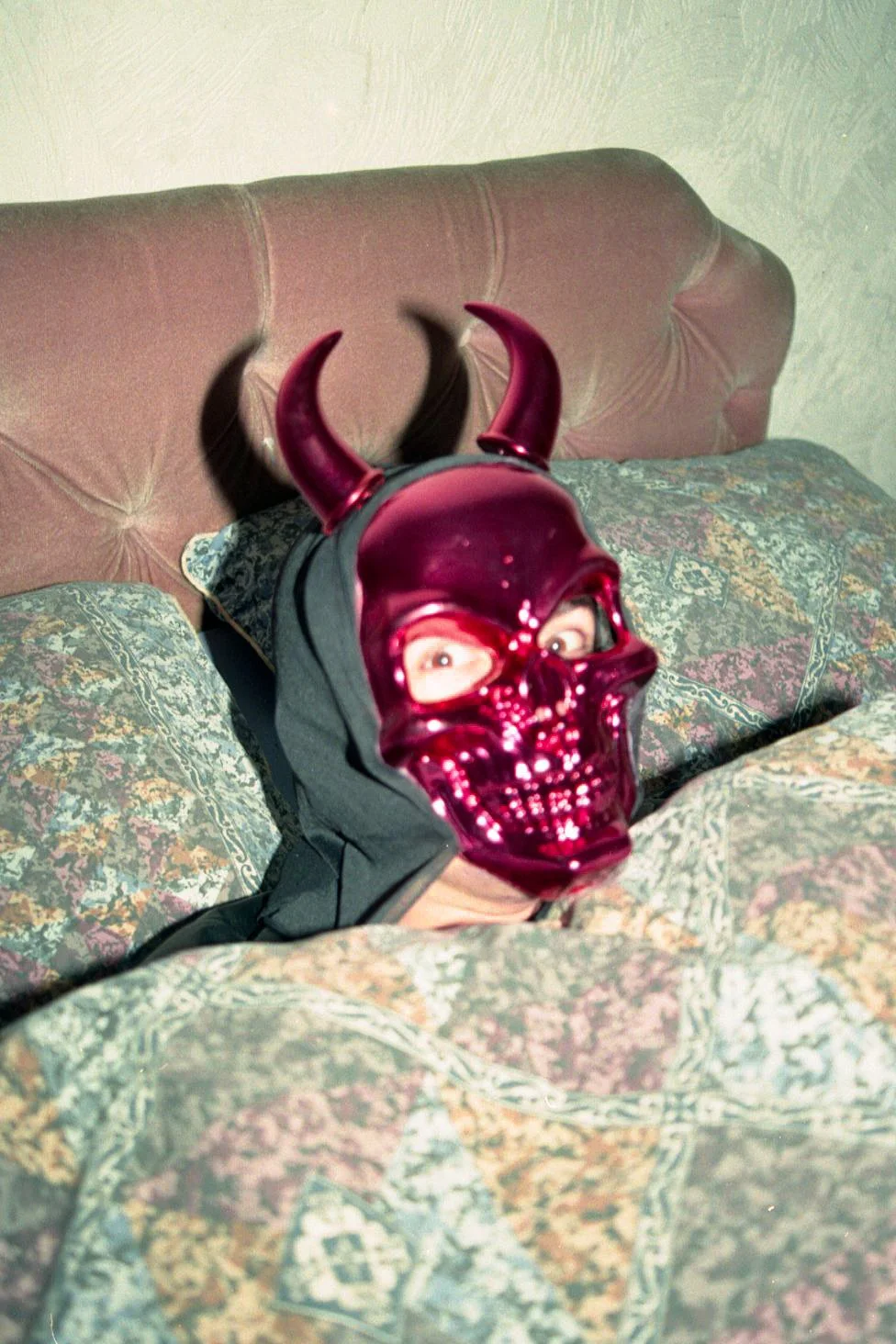 Person lying on a bed wearing a shiny purple skull mask with horn extensions.