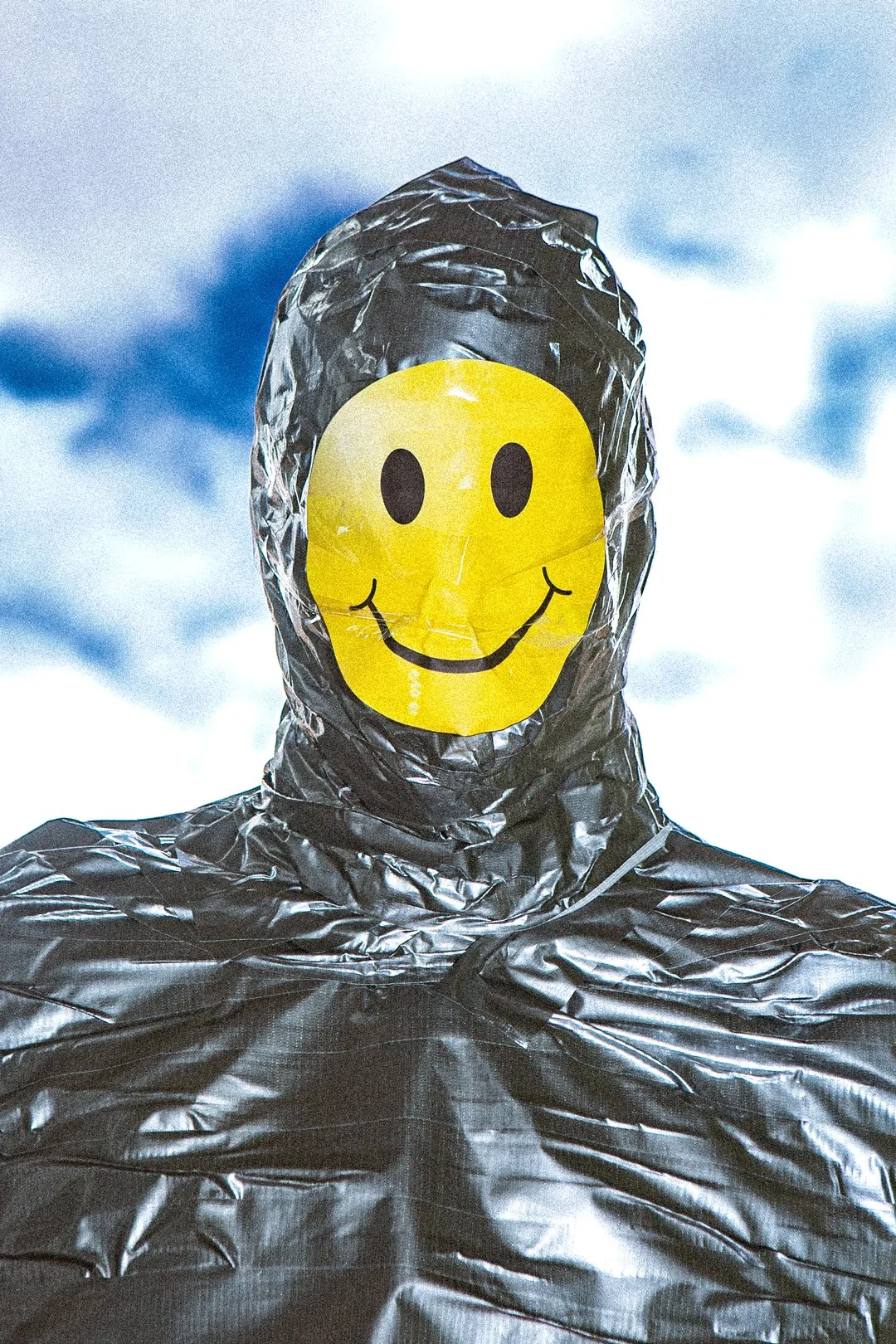 Person wearing a black, shiny, hooded suit with their face covered by a yellow smiley face mask showing a happy expression, against a cloudy sky background.