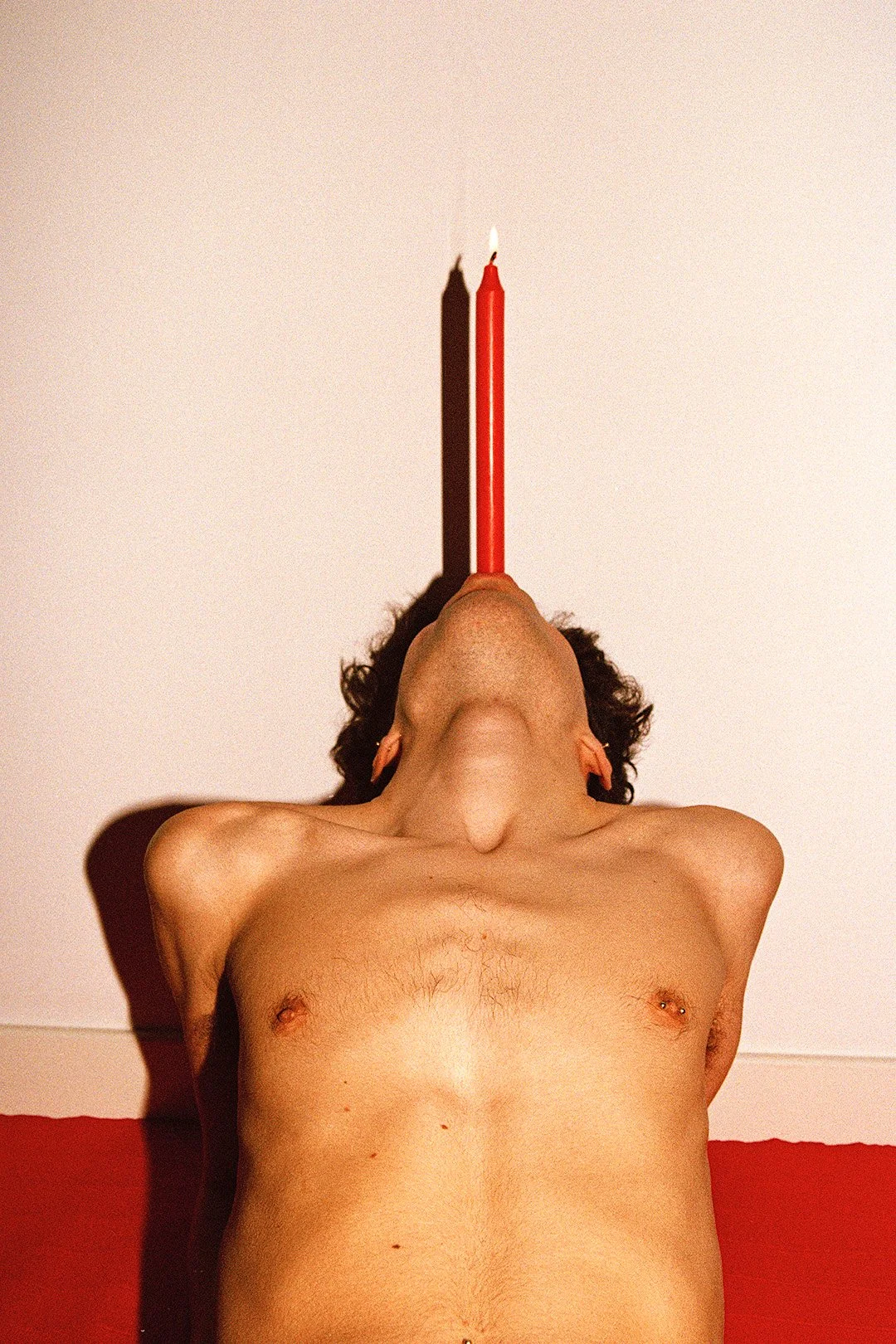 A topless woman lying on her back with eyes closed and arms behind her head, balancing a lit red candle on her nose against a white background.