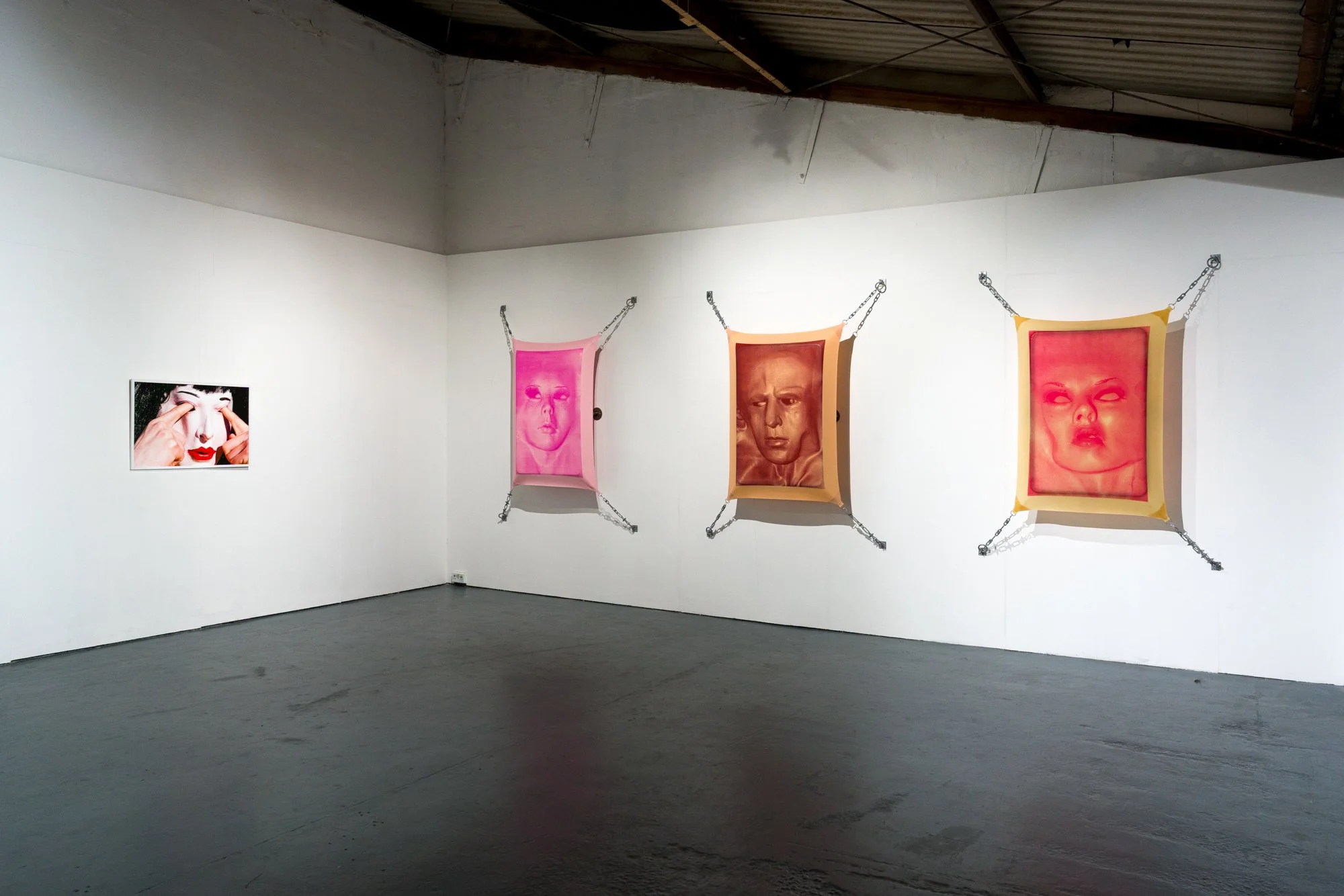 Art gallery with three portrait artworks hanging on a white wall, chain suspension display, and a photo of a woman with makeup on the left.