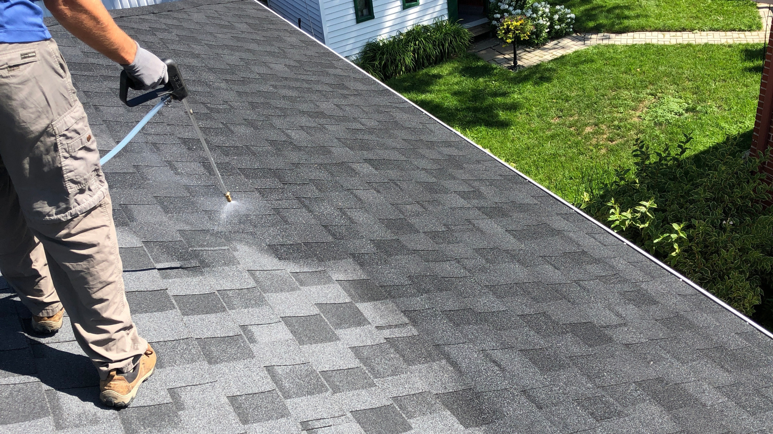 A person using a pressure washer to clean a gray asphalt shingle roof with a garden and house visible in the background.