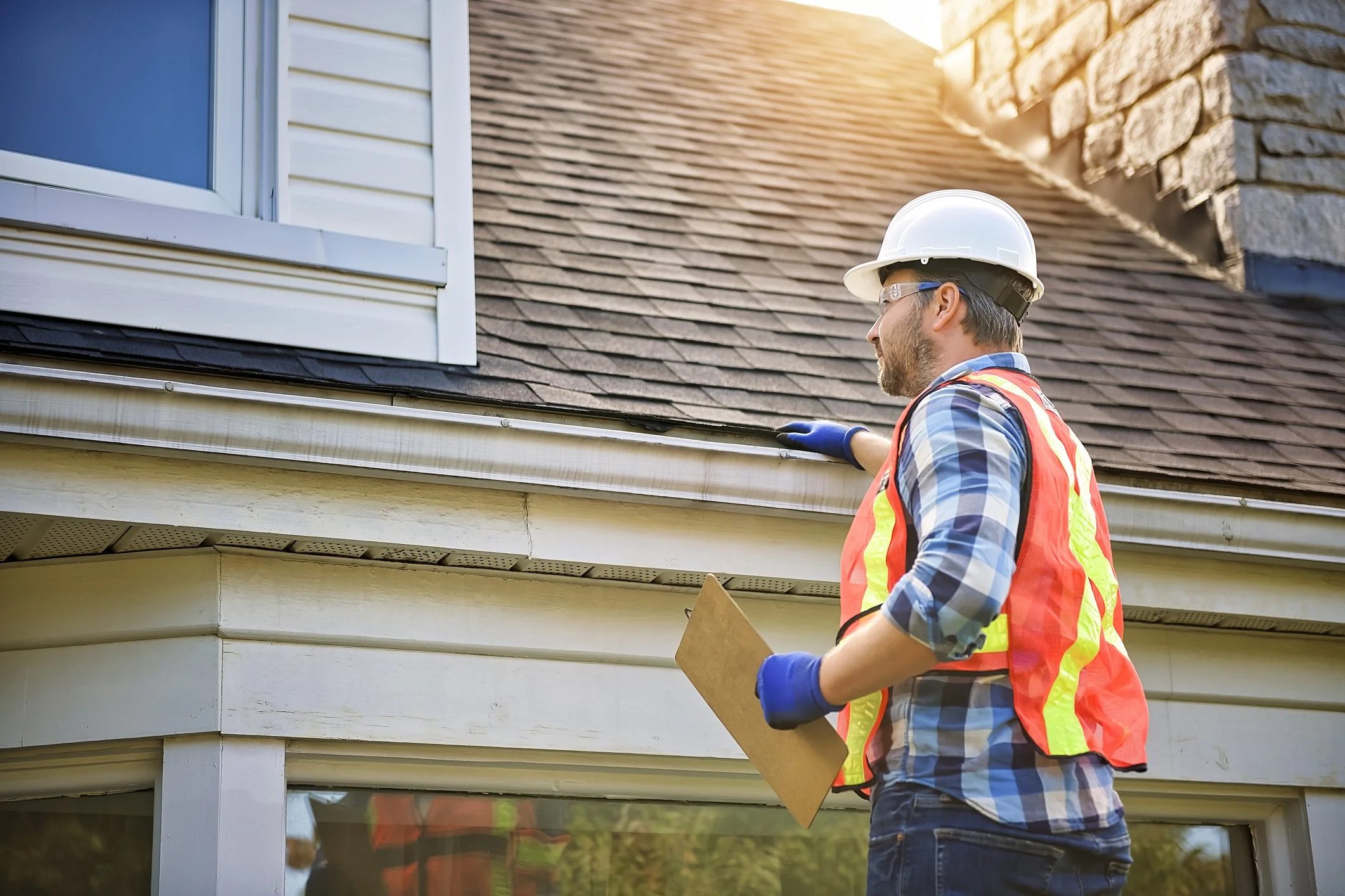 A man wearing a white hard hat, safety goggles, an orange and yellow safety vest, and gloves inspecting a house's exterior siding and soffit.