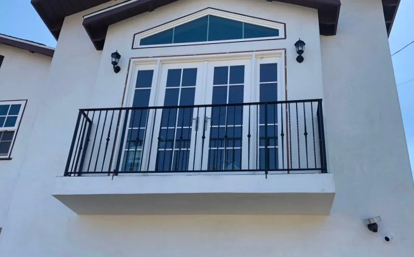 Front view of a house with a balcony, white doors with multiple glass panes, black railing, two wall-mounted lanterns, and a small window to the left.