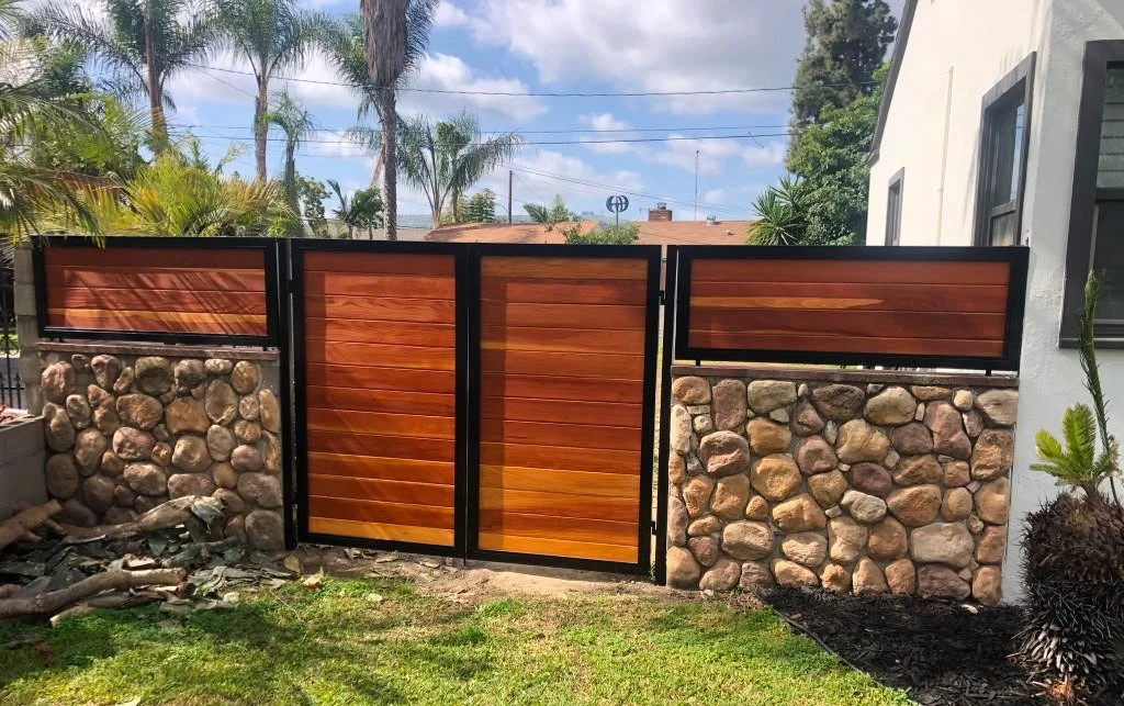 Wood and stone fence gate in a backyard with palm trees and a house visible behind.