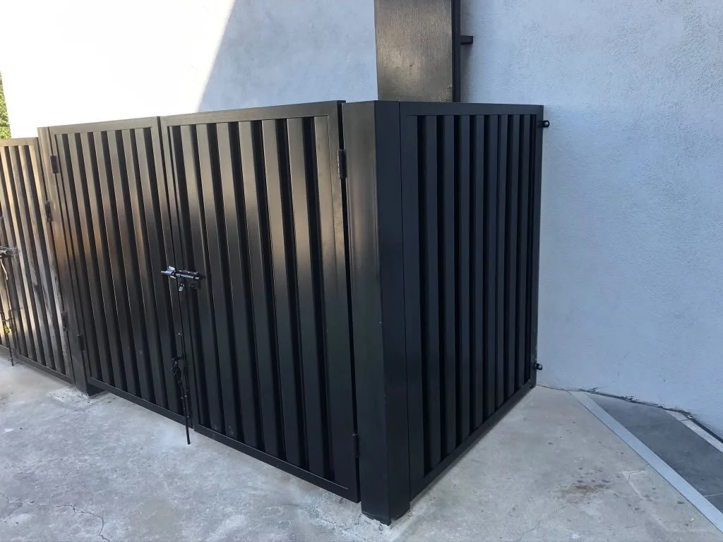 Black metal storage enclosure with vertical slats, located next to a white wall and concrete floor.