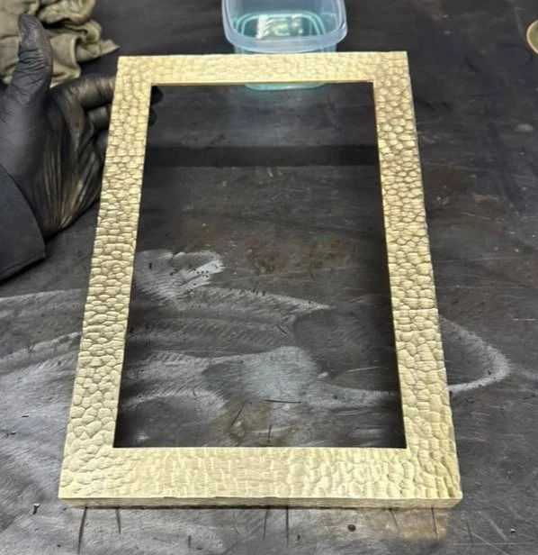 A rectangular picture frame with a textured, stone-like surface, placed on a dark wooden workbench.