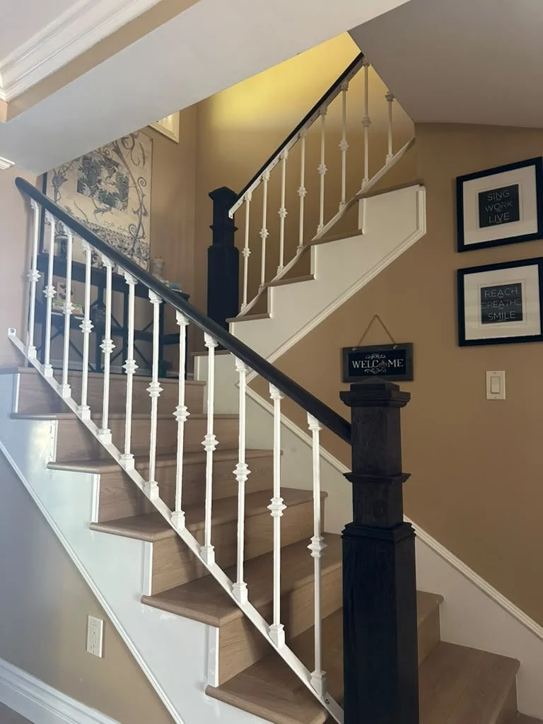 Indoor staircase with wooden steps, white railing with decorative spindles, black handrail, and beige walls decorated with framed art and a small welcome sign.