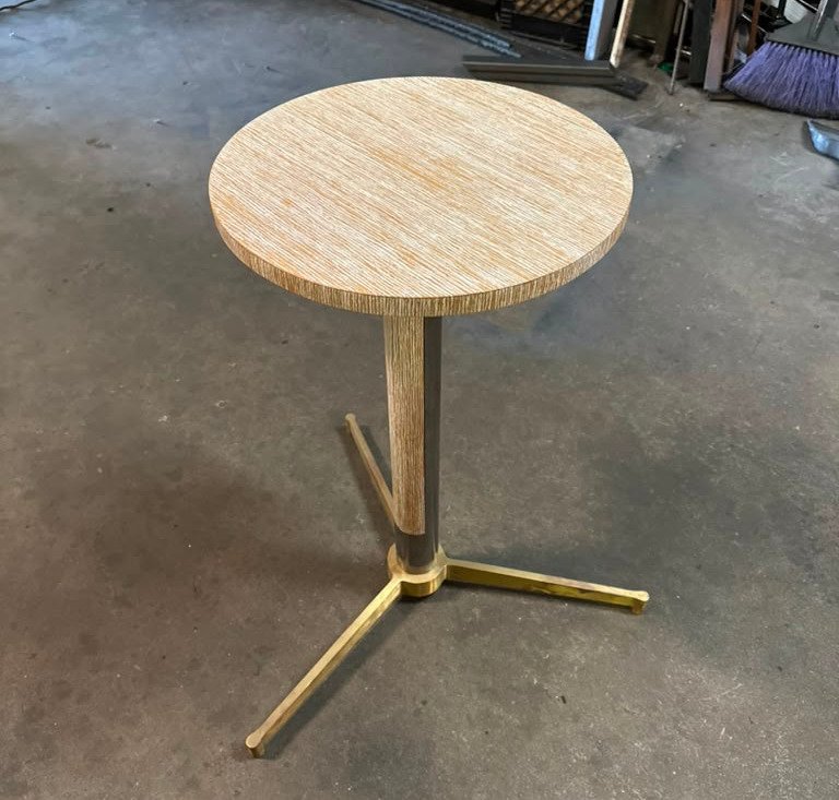 A round wooden table with a metal and wood base standing on a concrete floor in a workshop.