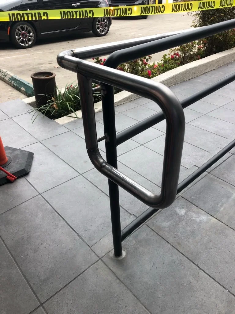 A metal railing on a sidewalk with a square opening in the middle. Behind it, there is yellow caution tape, a parked car, some plants, and flowers.
