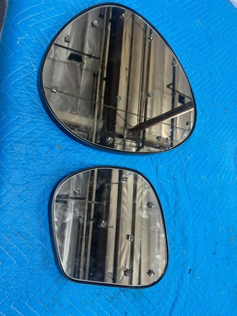 Two car side mirror glass pieces with mounting clips visible, resting on a blue padded surface. Reflection shows an industrial setting with metal beams and equipment.