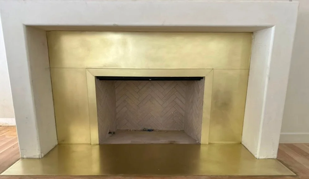 A fireplace with a white surround and a brass mantle and hearth, with a tiled interior in a herringbone pattern.