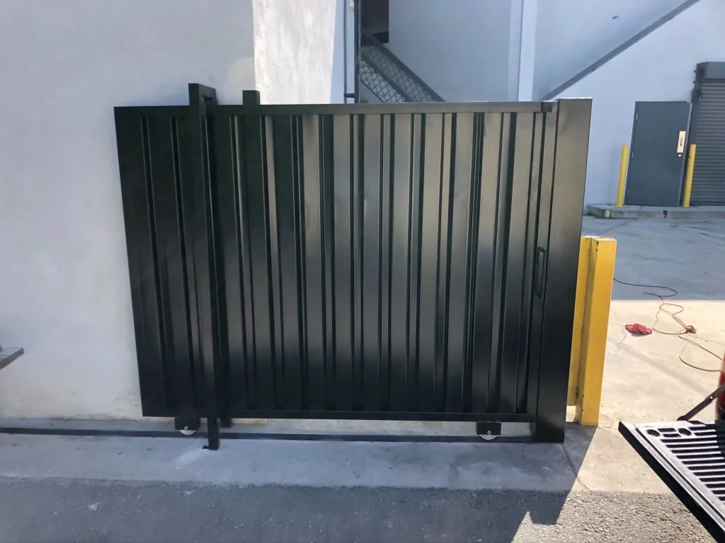 Black metal sliding gate installed against a white wall in an industrial or commercial area, with yellow safety posts and a chain link fence visible in the background.