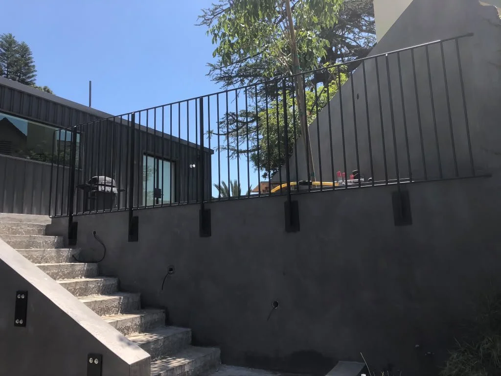An outdoor staircase leading up to a patio with a black metal railing, a charcoal grill, and construction tools in an area with trees and a modern building.