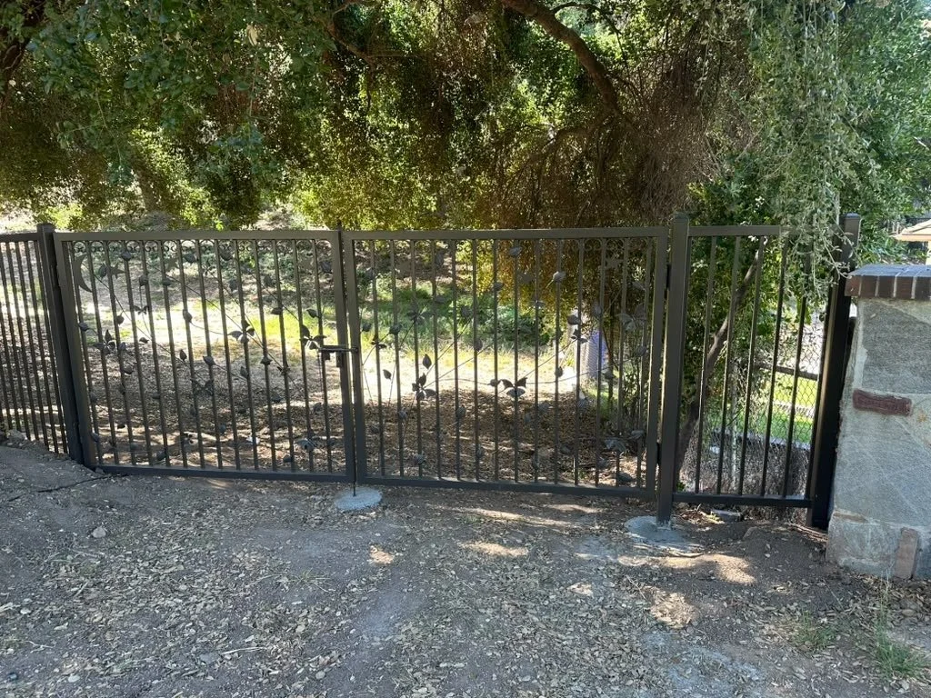 A black metal fence with a gate in front of a leafy yard, trees, and some sunlight.