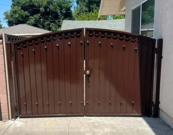 A double metal gate painted brown with decorative scrollwork at the top, installed at the entrance to a driveway.