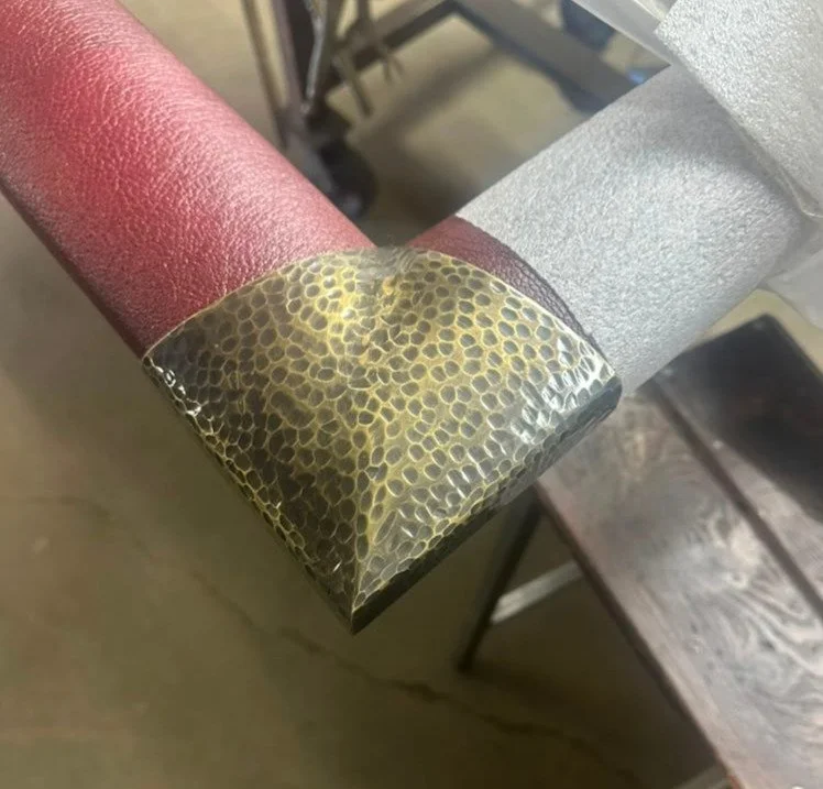 Close-up of a corner of a metal object with a yellow and black textured protective tape or coating, adjacent to a painted pink surface, with a blurred wooden stool in the background.