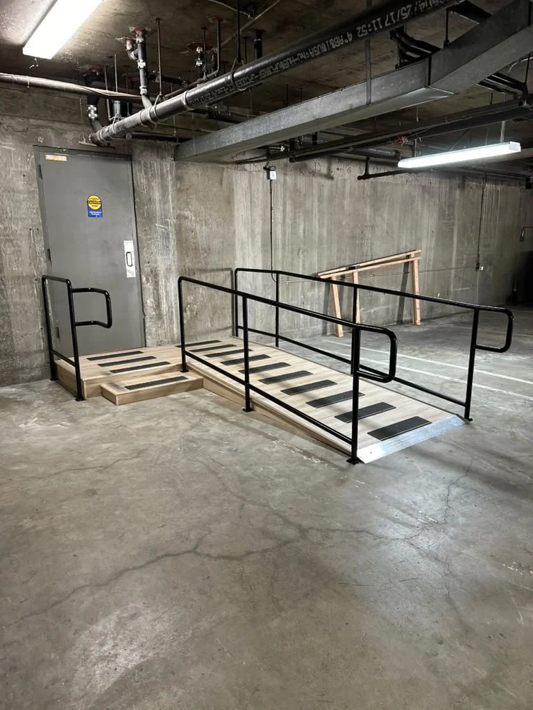 An indoor accessibility ramp with black railings, made of wood and metal, in an industrial setting with concrete walls and ceiling pipes.