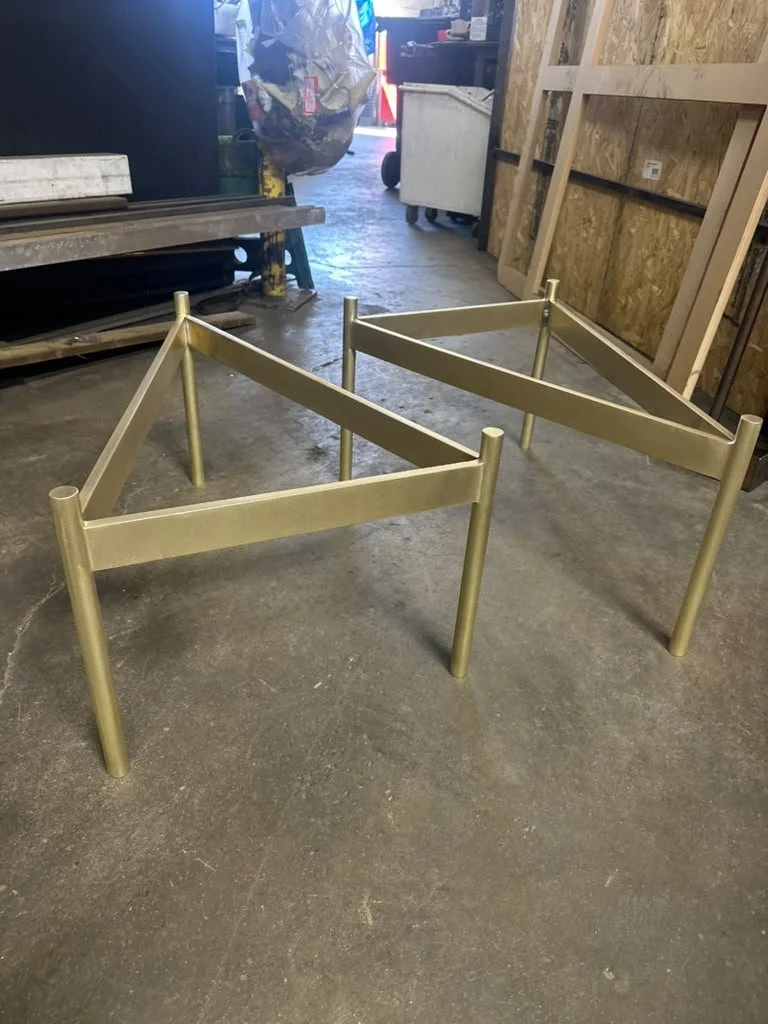 Two metallic bed frames without mattresses on a concrete shop floor, with workshop supplies and wooden panels in the background.