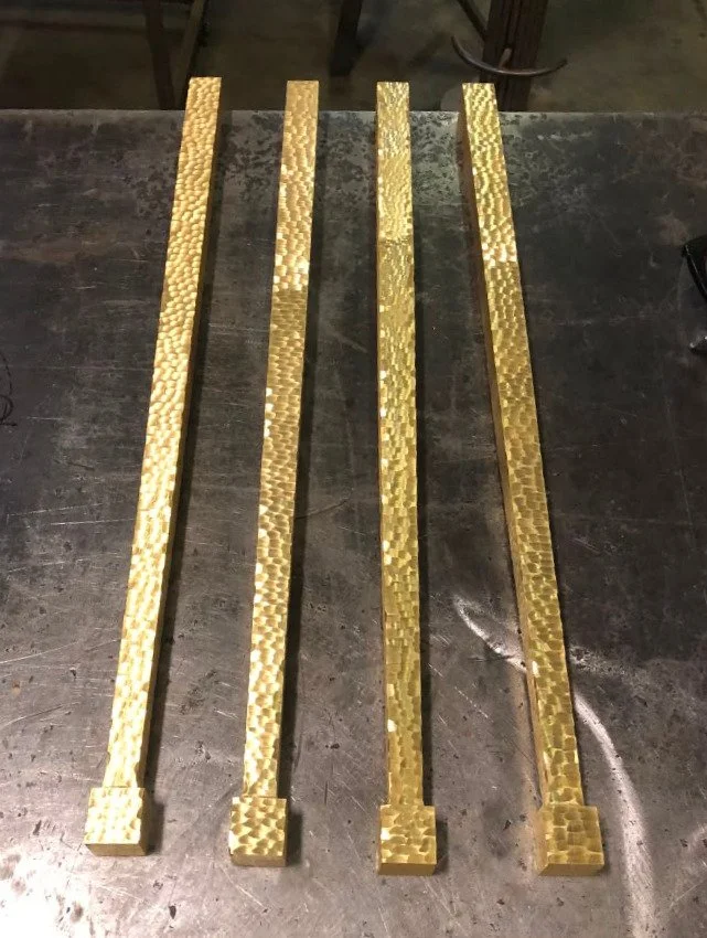 Four elongated gold-colored metal bars with textured, hammered surfaces, resting on a dark wooden surface.