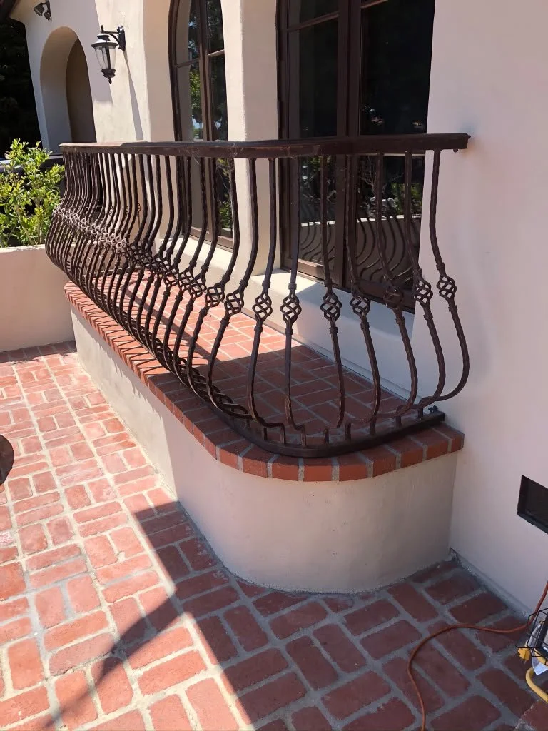 A small brick balcony with wrought iron railing outside a house with white stucco walls and large windows.
