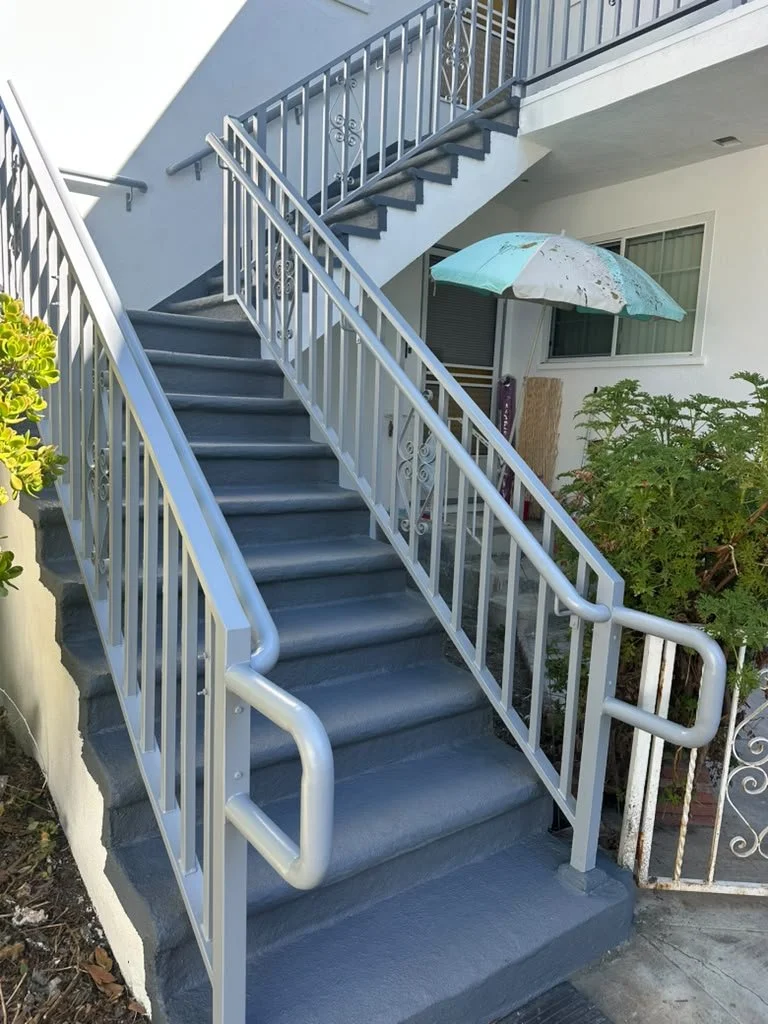 Exterior metal staircase with handrails leading up to a second-floor balcony of a white building. An umbrella is attached to a railing on the balcony, and there are plants near the stairs.