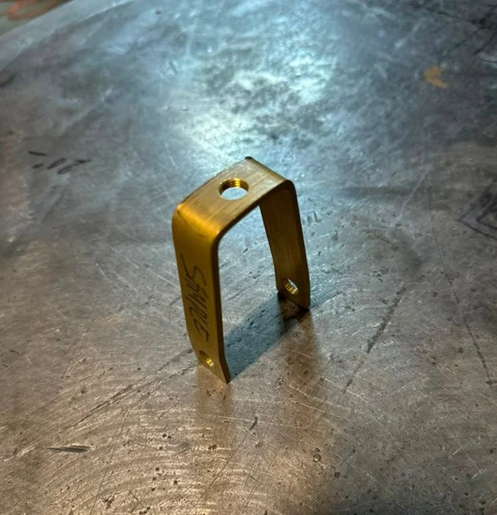 A small, gold-colored, metallic component with a curved shape and threaded holes on top and bottom, resting on a scratched metallic surface.
