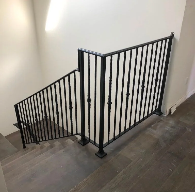 Black metal staircase railing along a staircase landing, with wooden stairs and a (partially visible) off-white wall.
