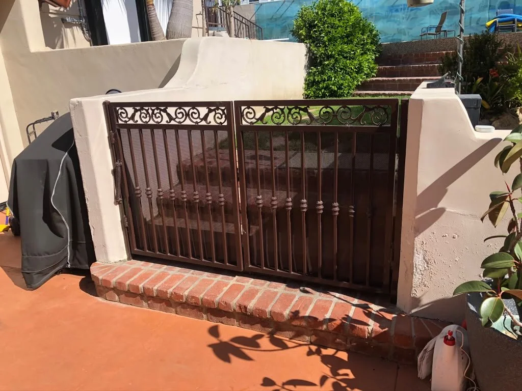 A small decorative metal gate with scrollwork on top, separating a brick patio from an outdoor staircase, with a lush green shrub in the background and various plants around.