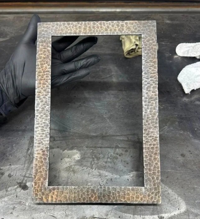 A gloved hand holding a small rectangular frame with a textured, stone-like border on a dark work surface, with cloth in the background.