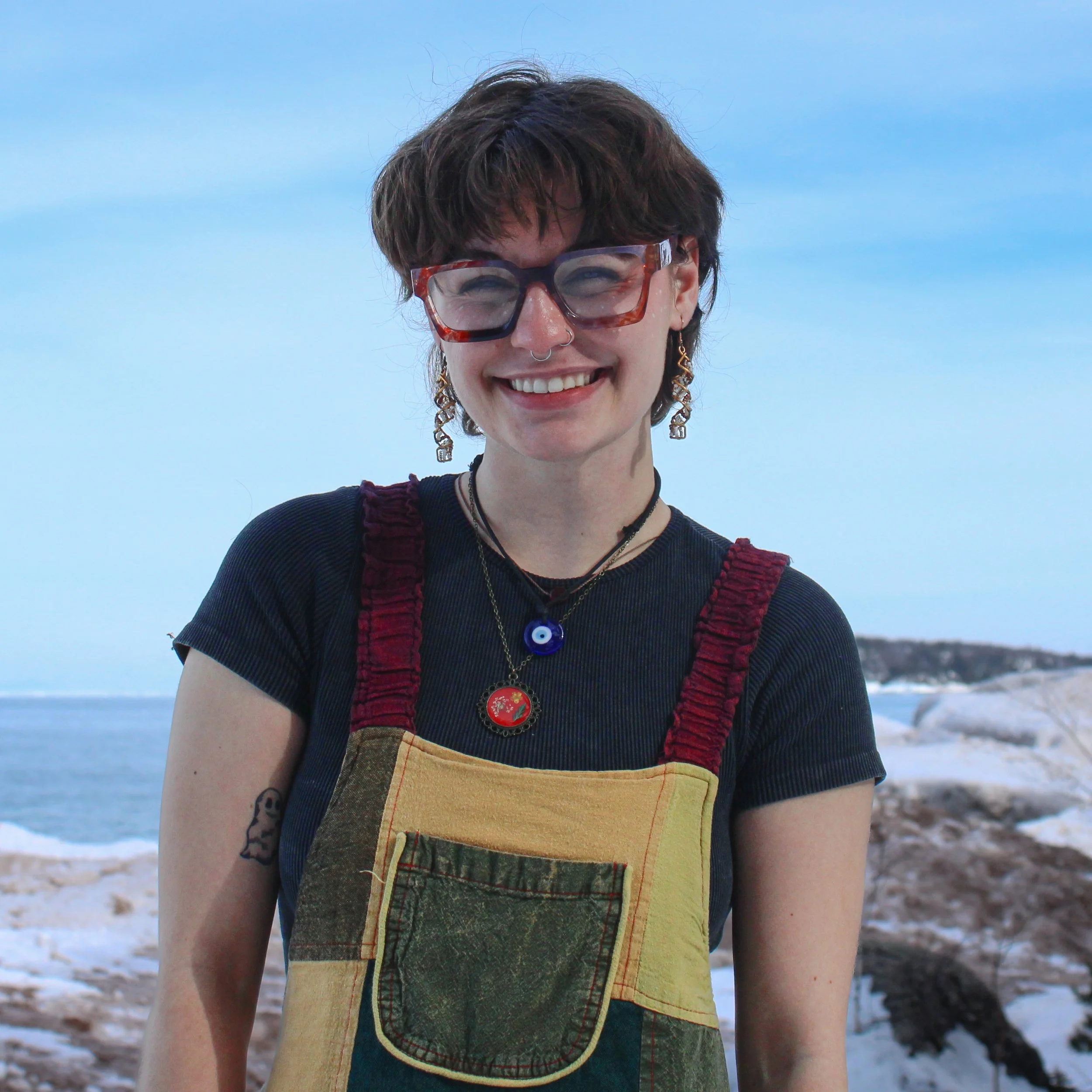 Young woman with short dark hair and glasses smiling outdoors near the water, wearing a patched apron and layered necklaces, with a snowy landscape in the background.