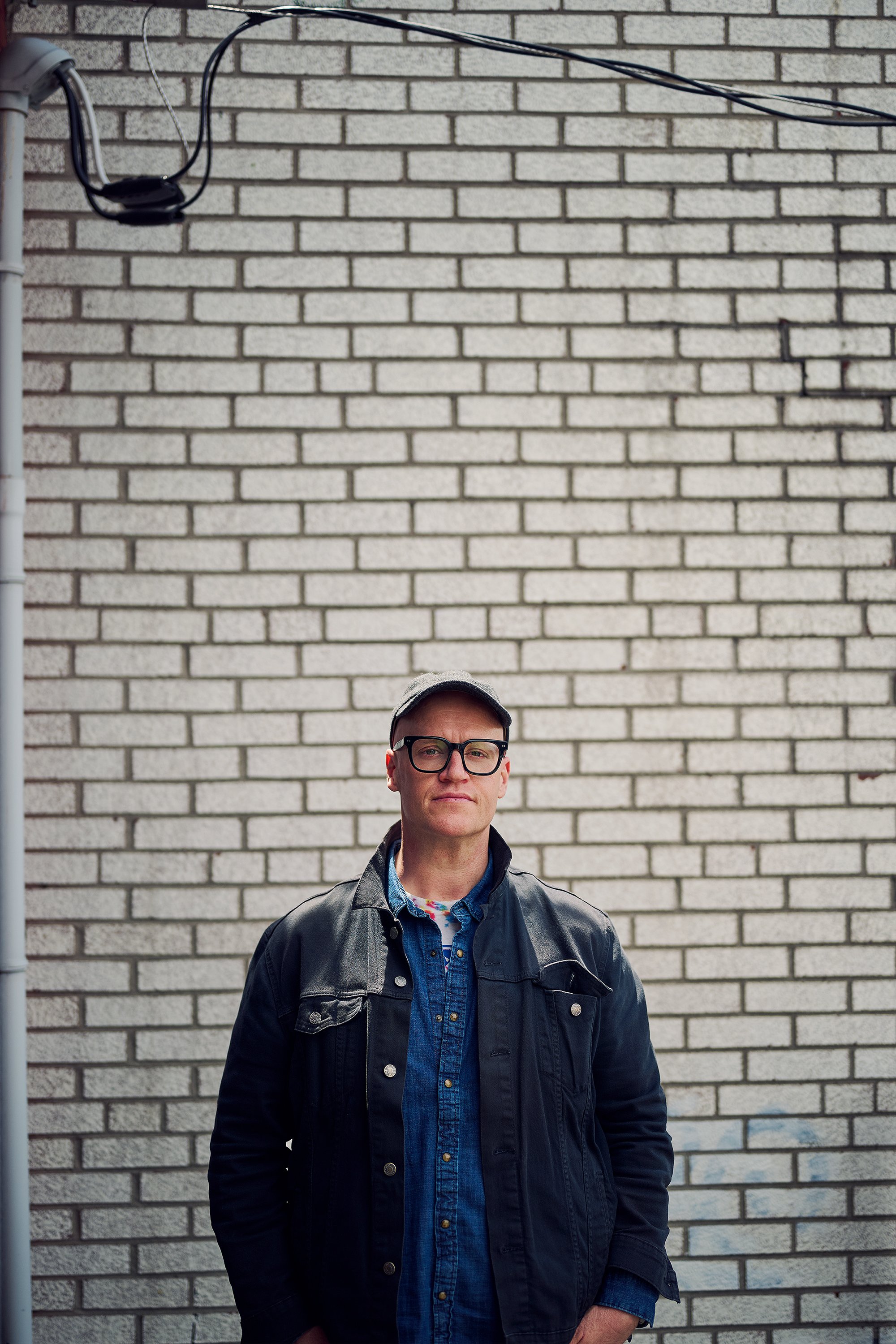 A man wearing glasses, a baseball cap, and a black jacket standing in front of a brick wall.