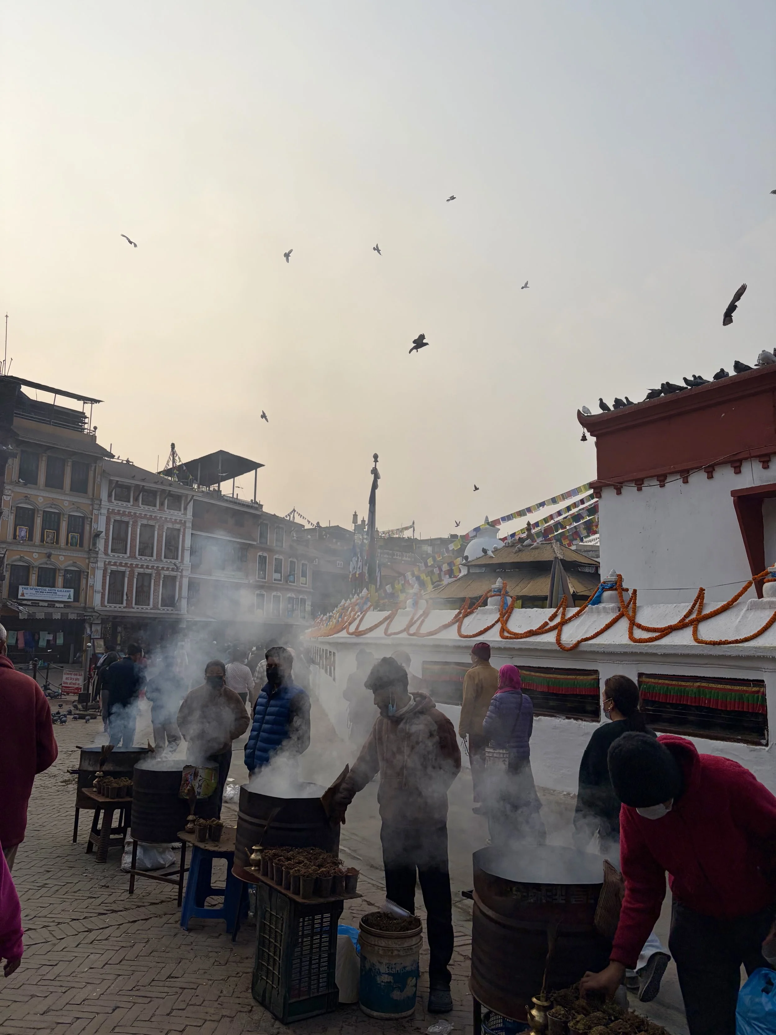 People gathered with smoke rising from fires or cooking stations, surrounded by buildings and decorative prayer flags, with birds flying in the sky.
