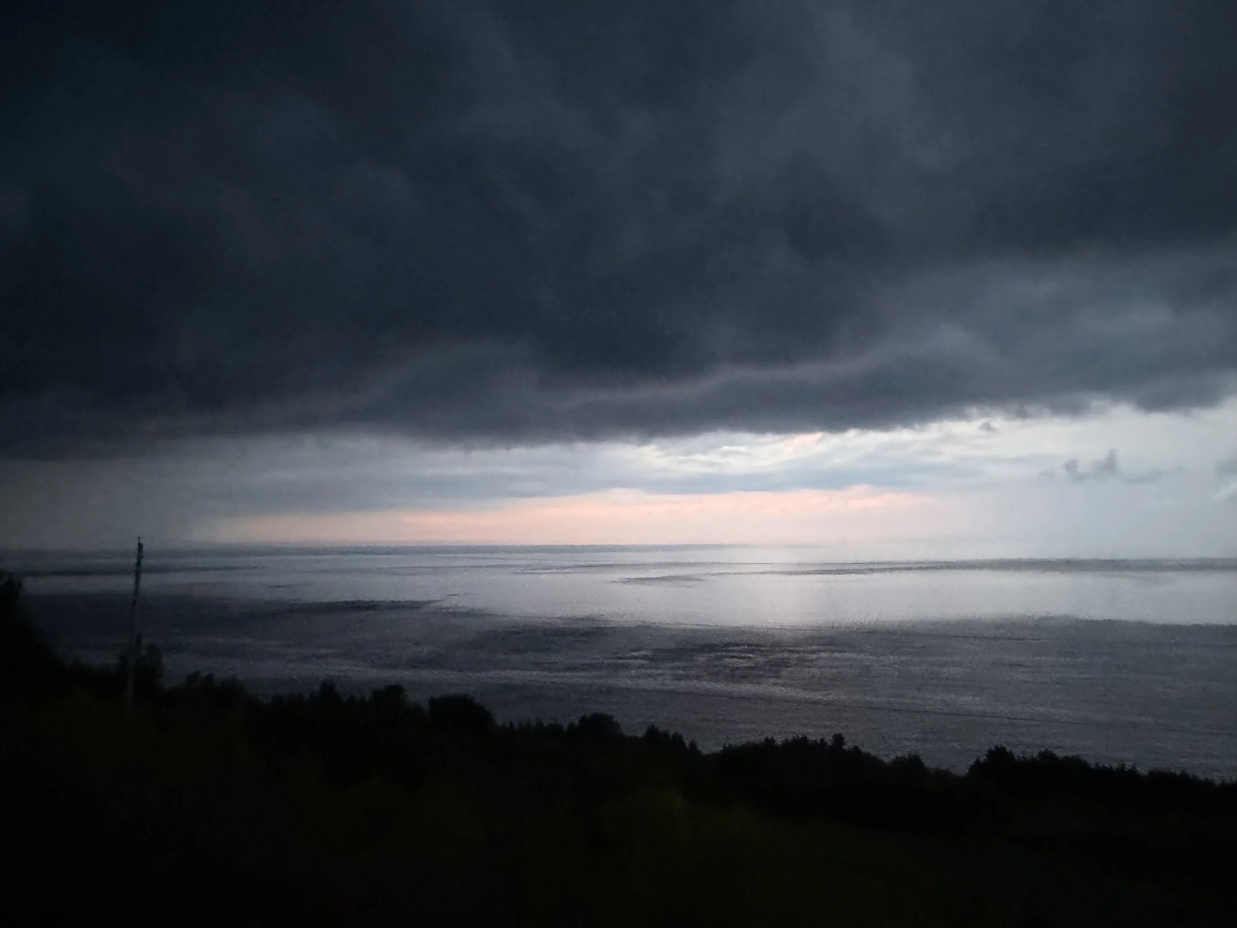 Dark storm clouds over a body of water at sunset, with silhouettes of trees and a small lighthouse in the distance.
