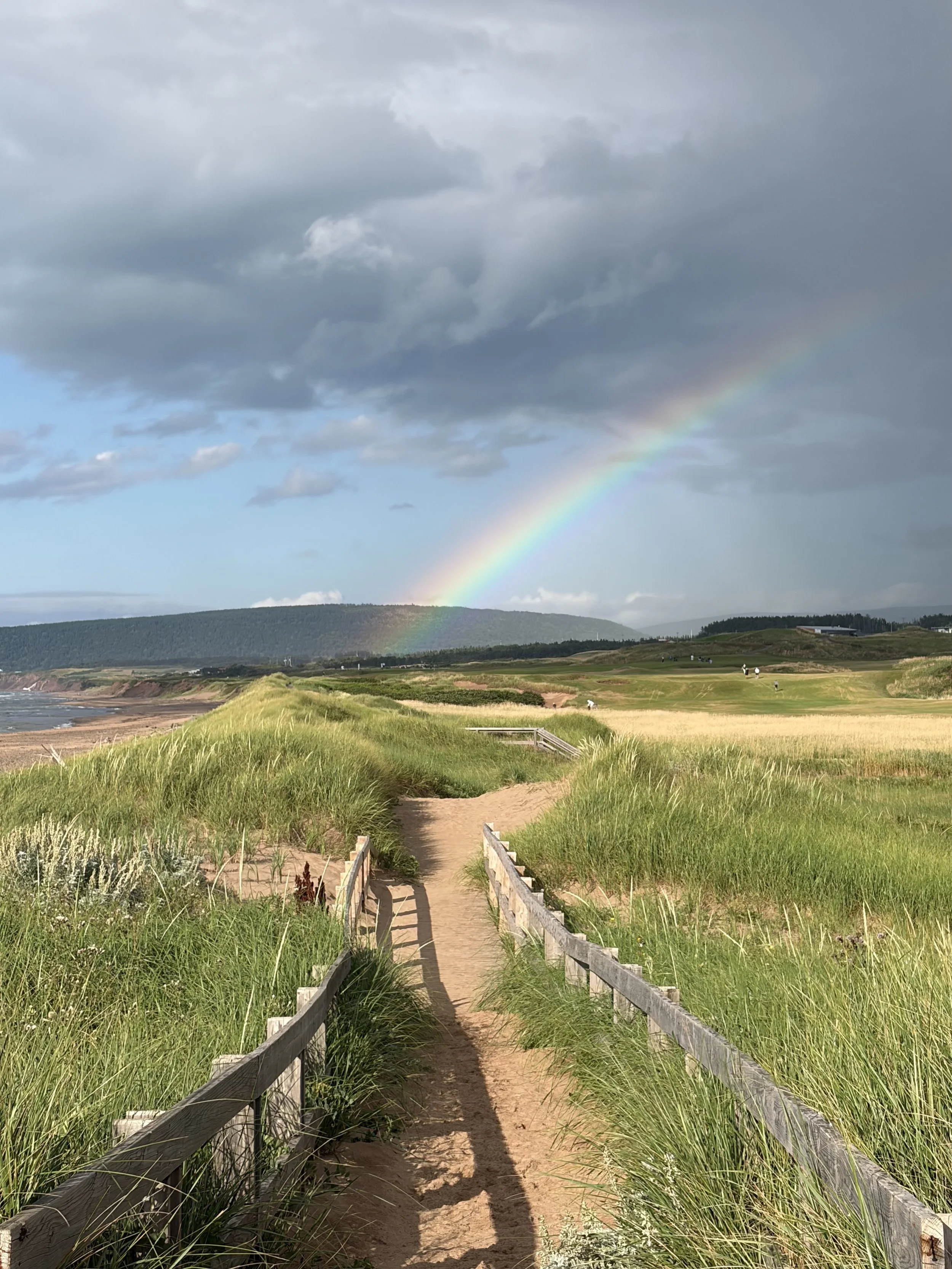 A sandy pathway with wooden railings on either side, leading through green grassy dunes towards a distant landscape. In the background, a cloudy sky with a faint rainbow arching across it.