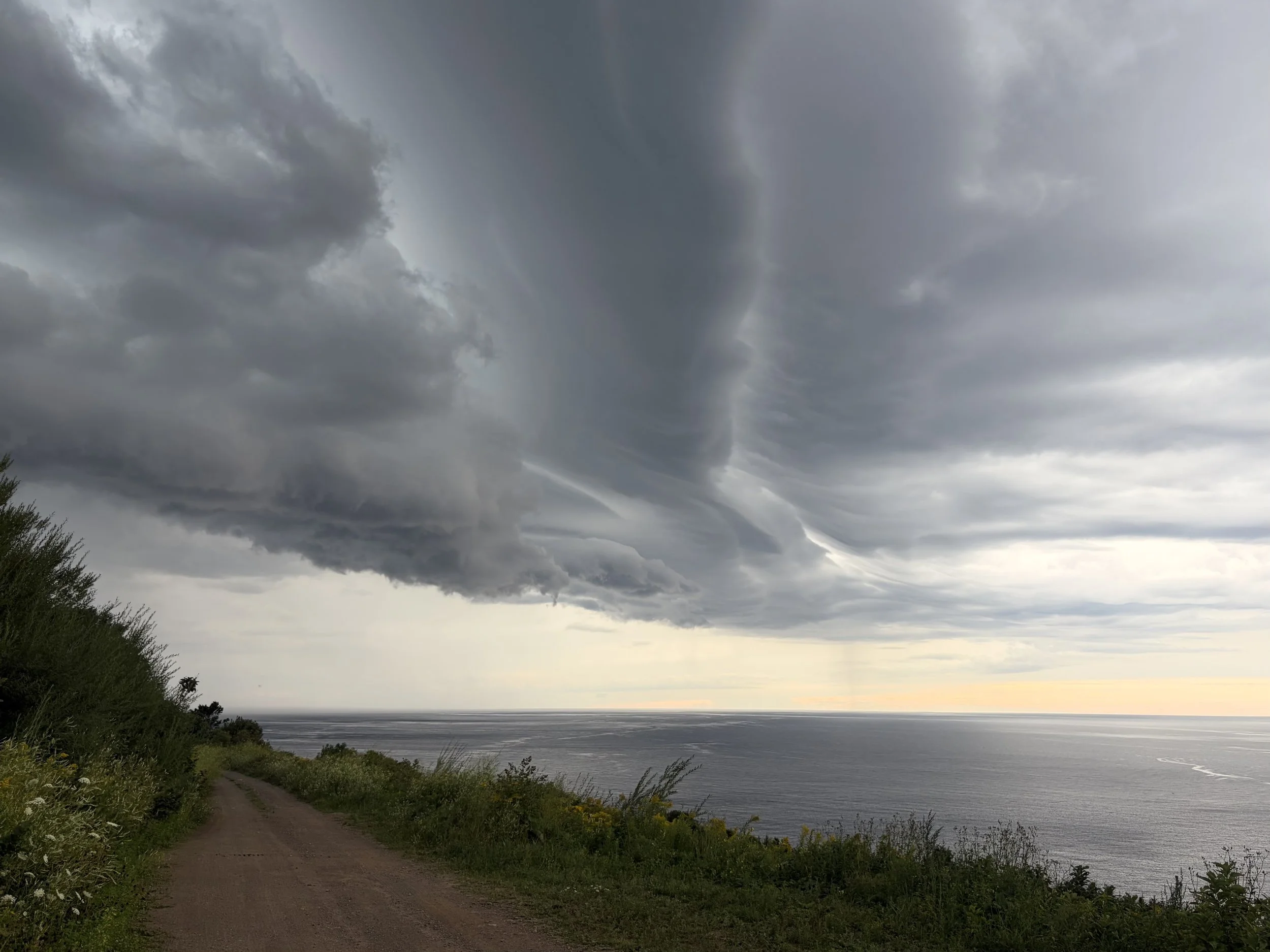 A dirt pathway runs along the coast with green bushes on each side. Dark storm clouds fill the sky above the ocean, which is visible in the background near the horizon. The ocean appears calm with a few small waves.