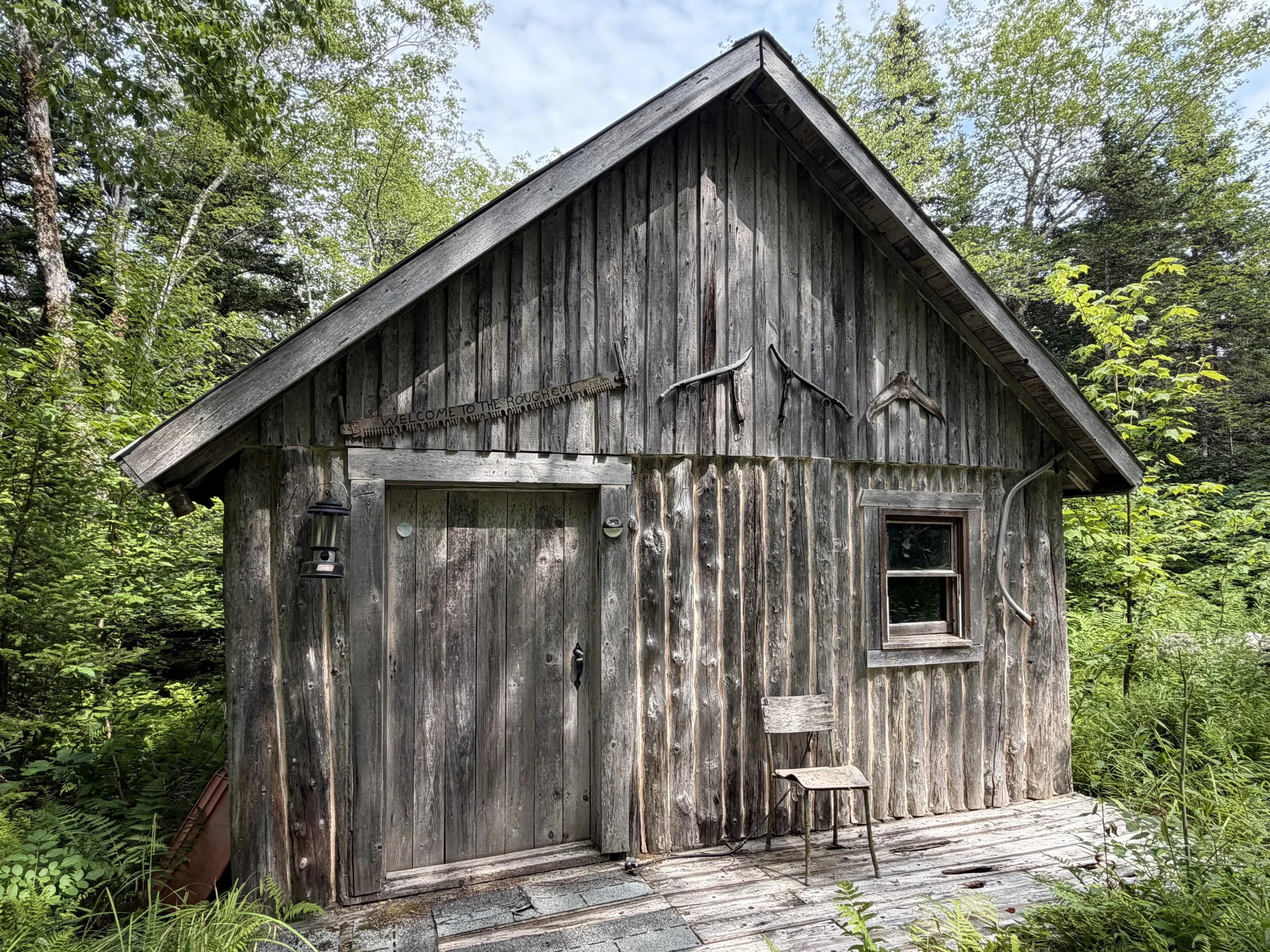 Old wooden cabin with a peaked roof, surrounded by green trees and foliage in a forest setting.