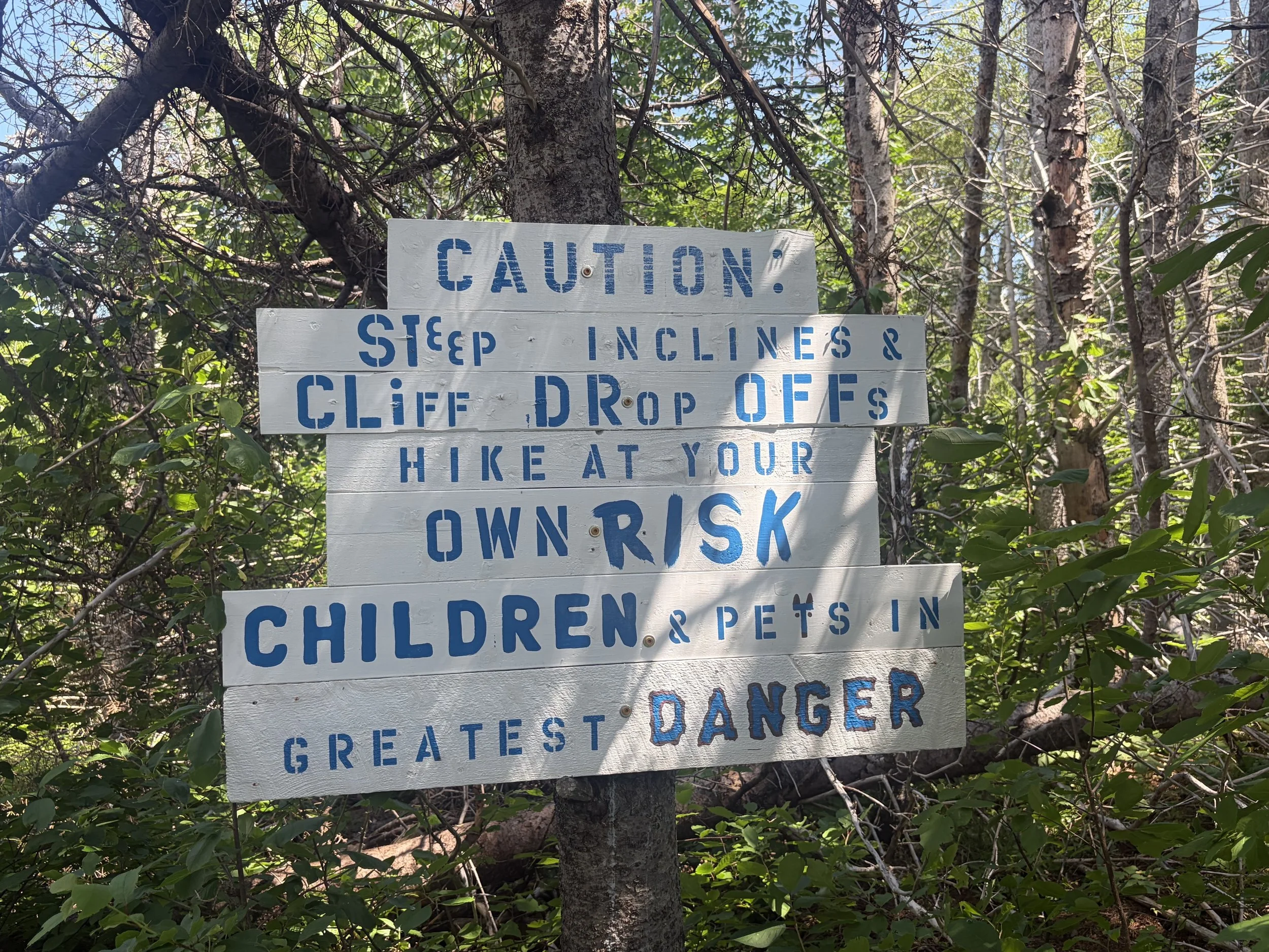 A warning sign attached to a tree in a forested area, cautioning about steep inclines, cliffs, and falling objects, and advising hikers to stay on the trail.