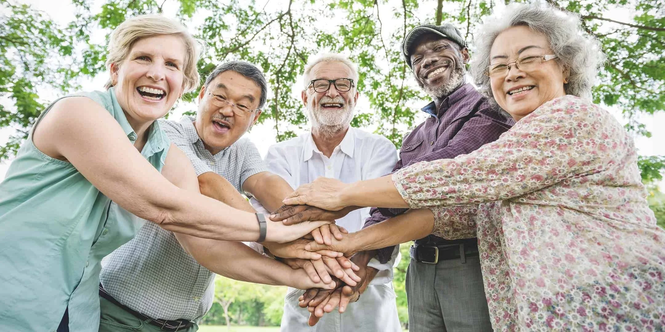 Six diverse elderly people standing outdoors in a circle with their hands stacked together, smiling and enjoying themselves.