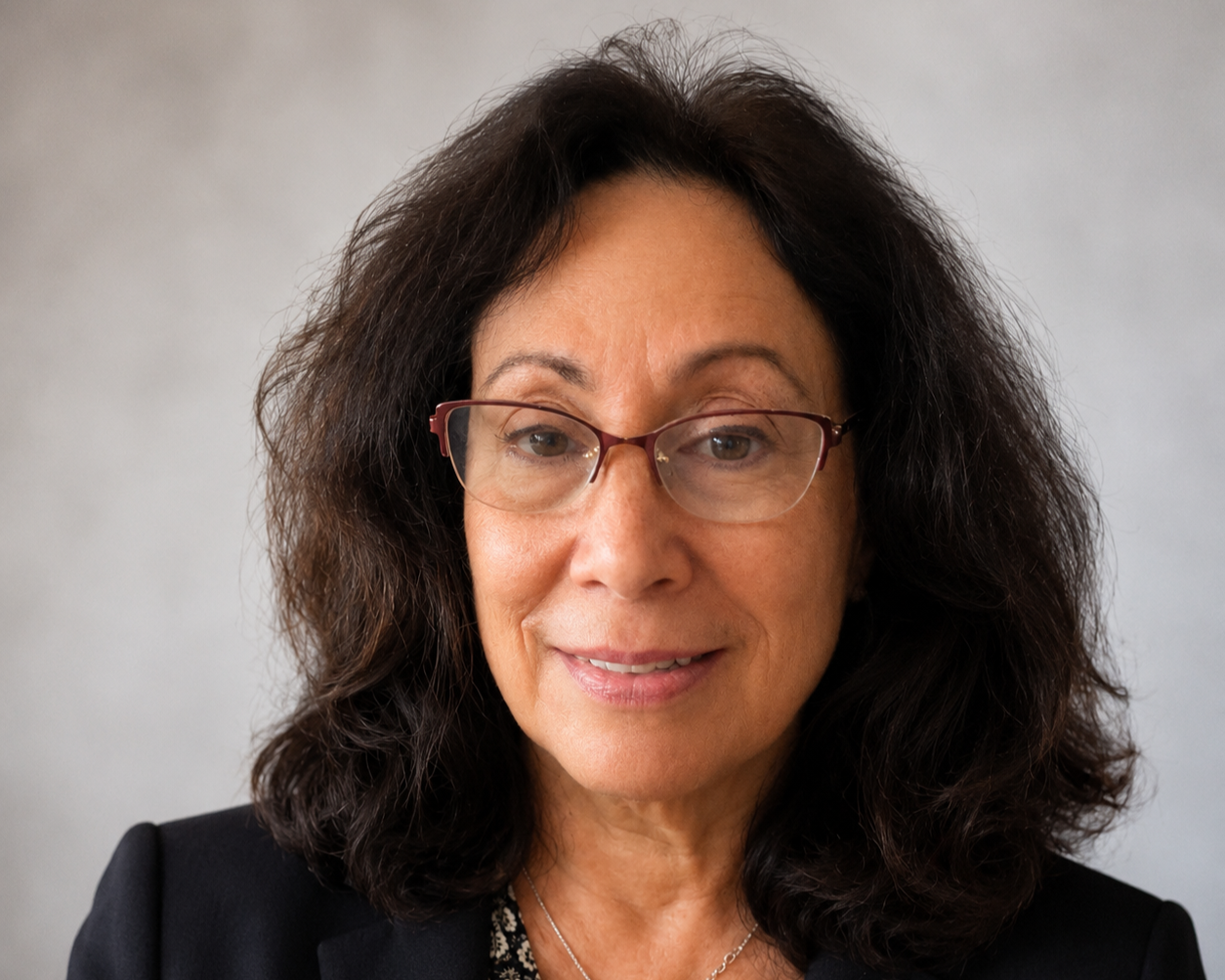 A woman with curly dark hair wearing glasses and a black blazer, smiling at the camera against a plain background.